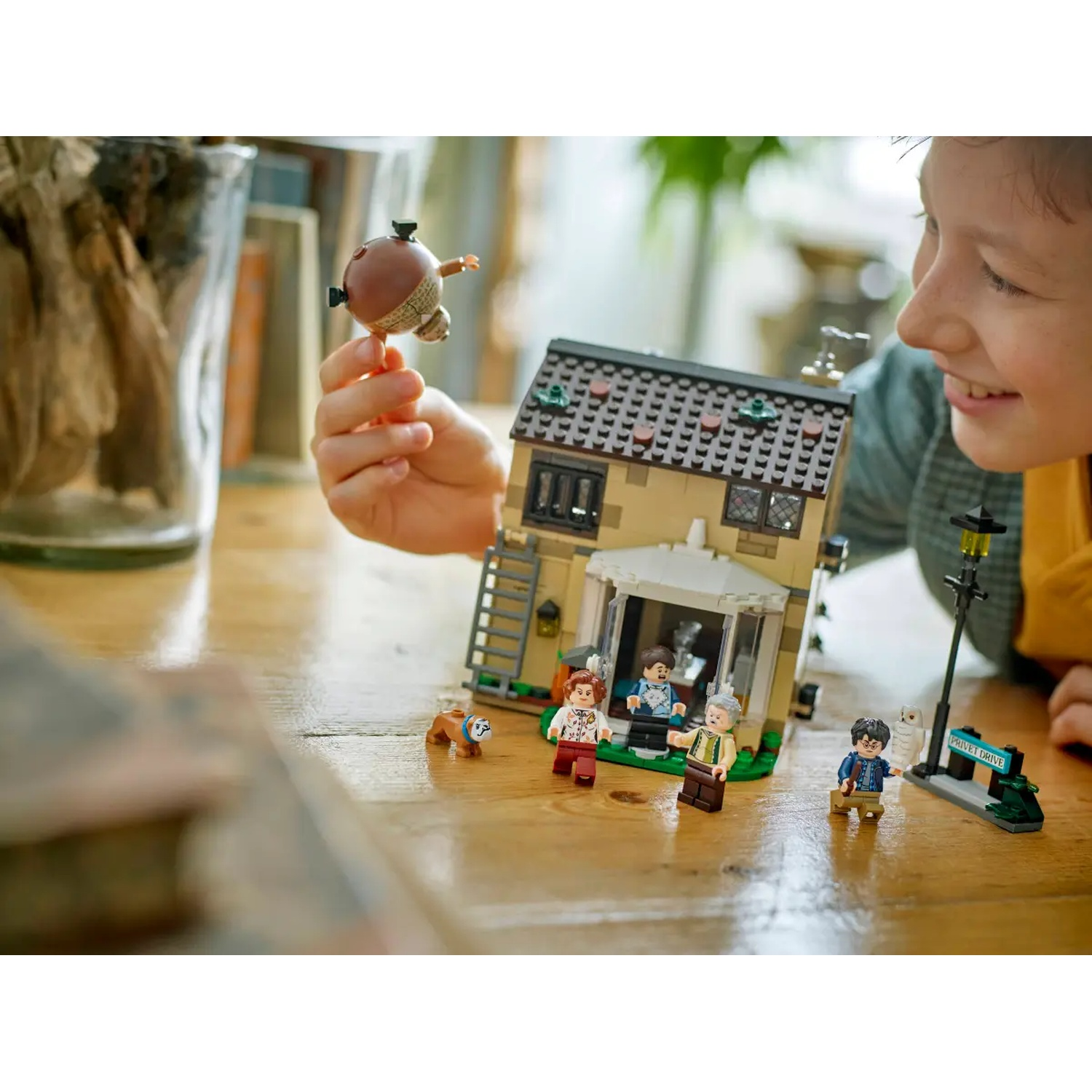A boy holds a toy. A Lego house with minifigures and a street sign are on a table. The boy smiles.