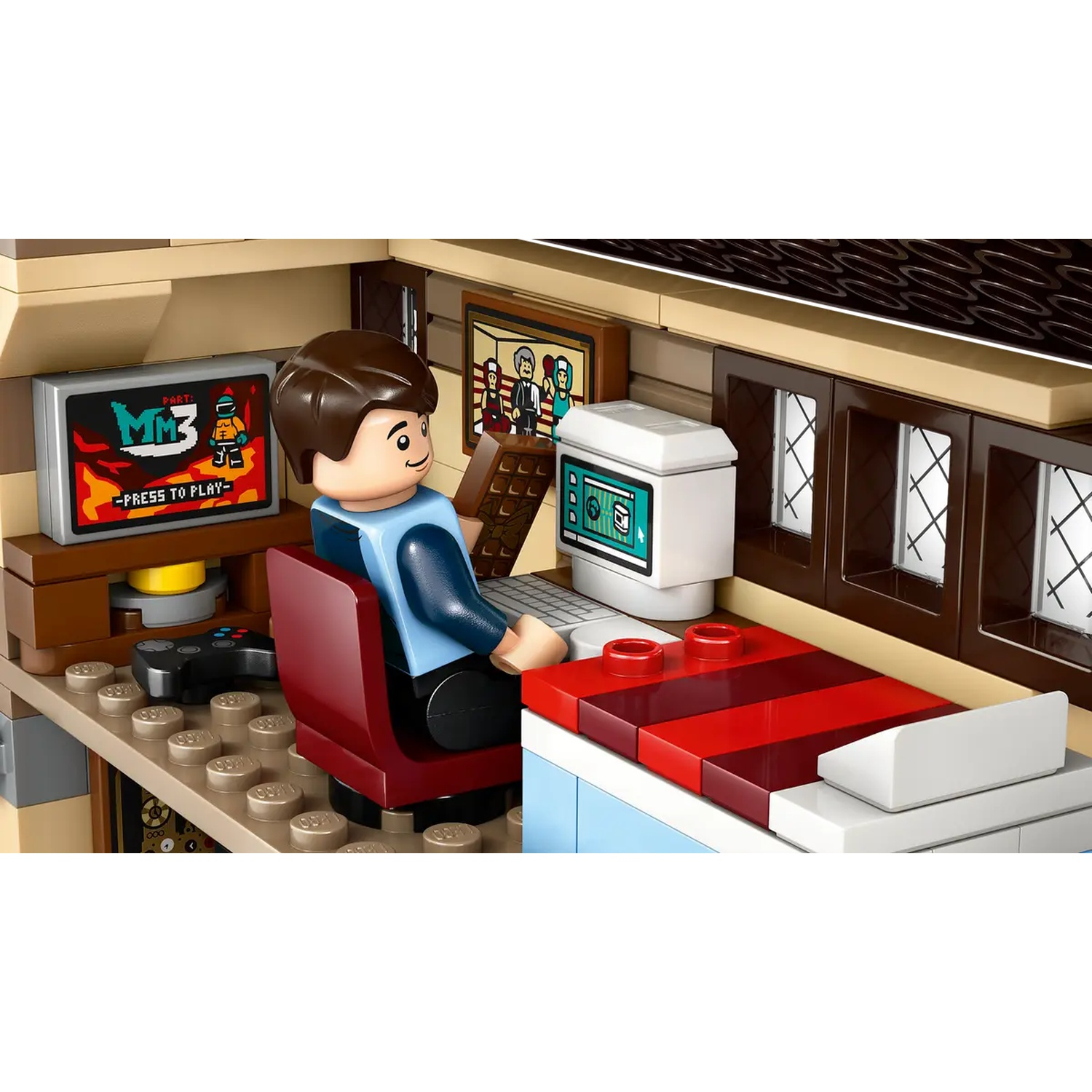 A Lego figure sits at a desk, playing a game. There's a computer, a screen showing a game, and a controller. A picture hangs on the wall.