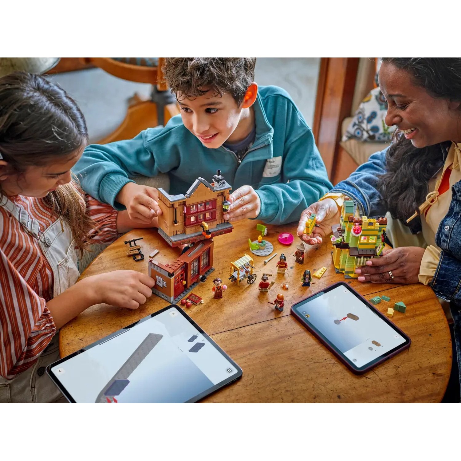 Three people build Lego sets around a table. Two tablets show augmented reality. Lego figures and buildings are scattered.