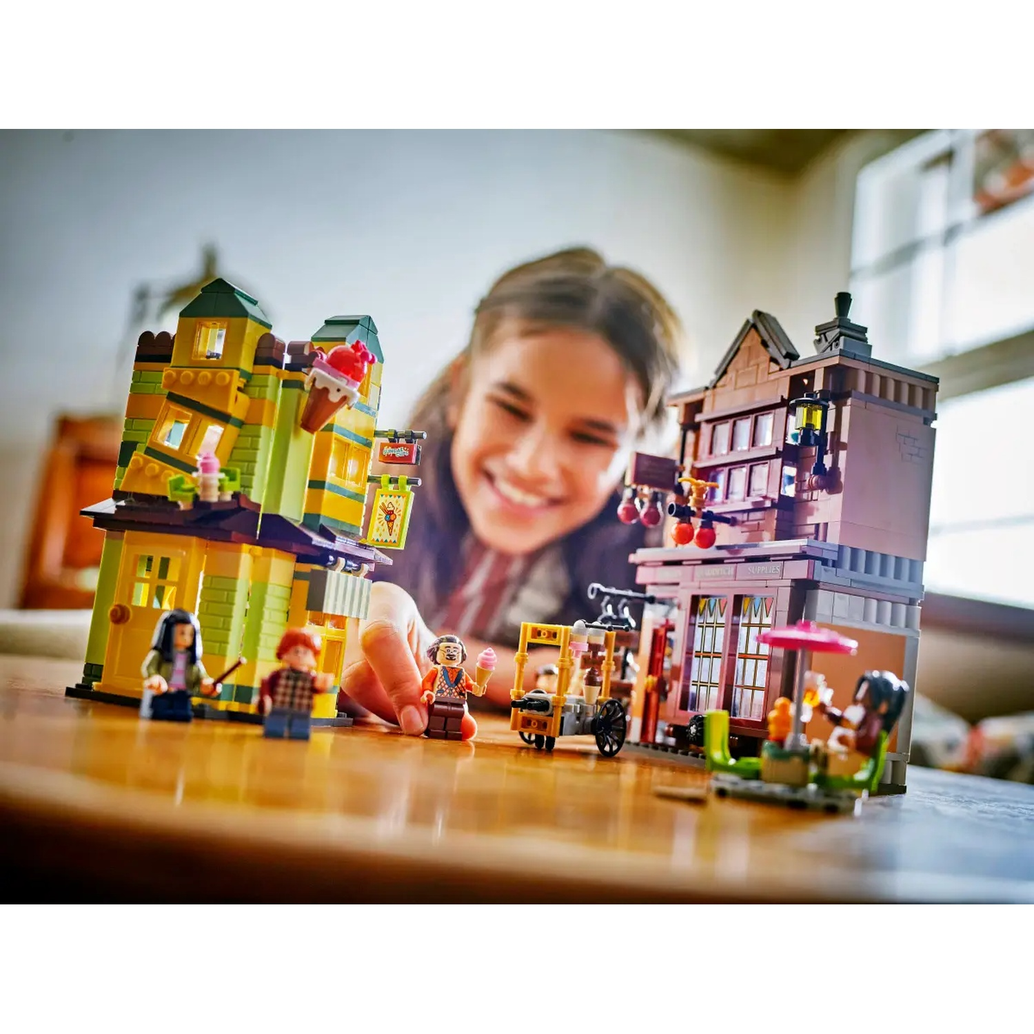 A child smiles at a Lego set with buildings, figures, and an ice cream cart. The set is on a wooden table.