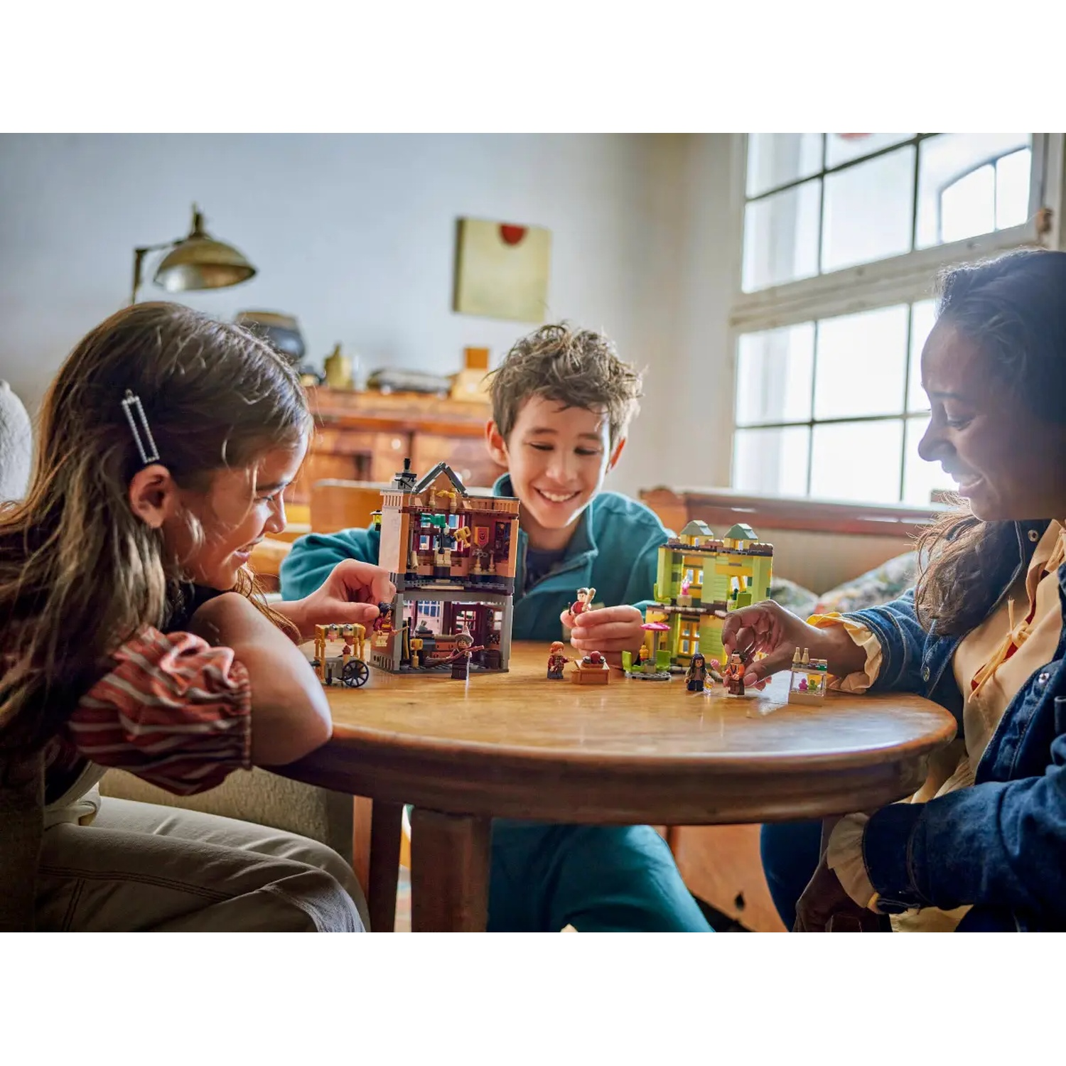 Three children play with Lego sets on a round table. One set is a brown haunted house, another is green. They smile.