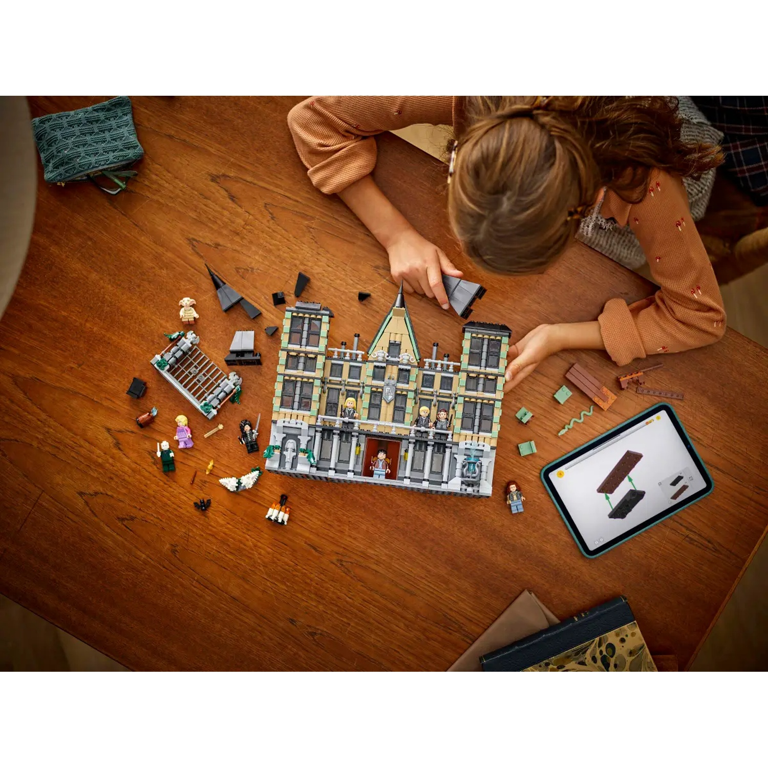 A girl assembling a Lego building on a wooden table. Lego pieces and figures are scattered around. A tablet shows instructions.
