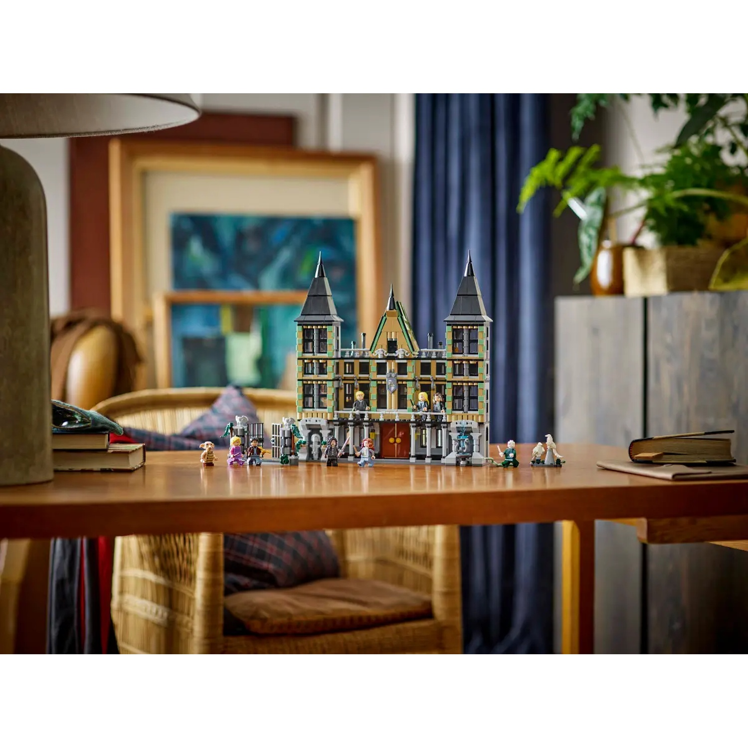 A Lego castle with mini figures on a wooden table. A lamp, paintings, plants, and a chair are in the background.