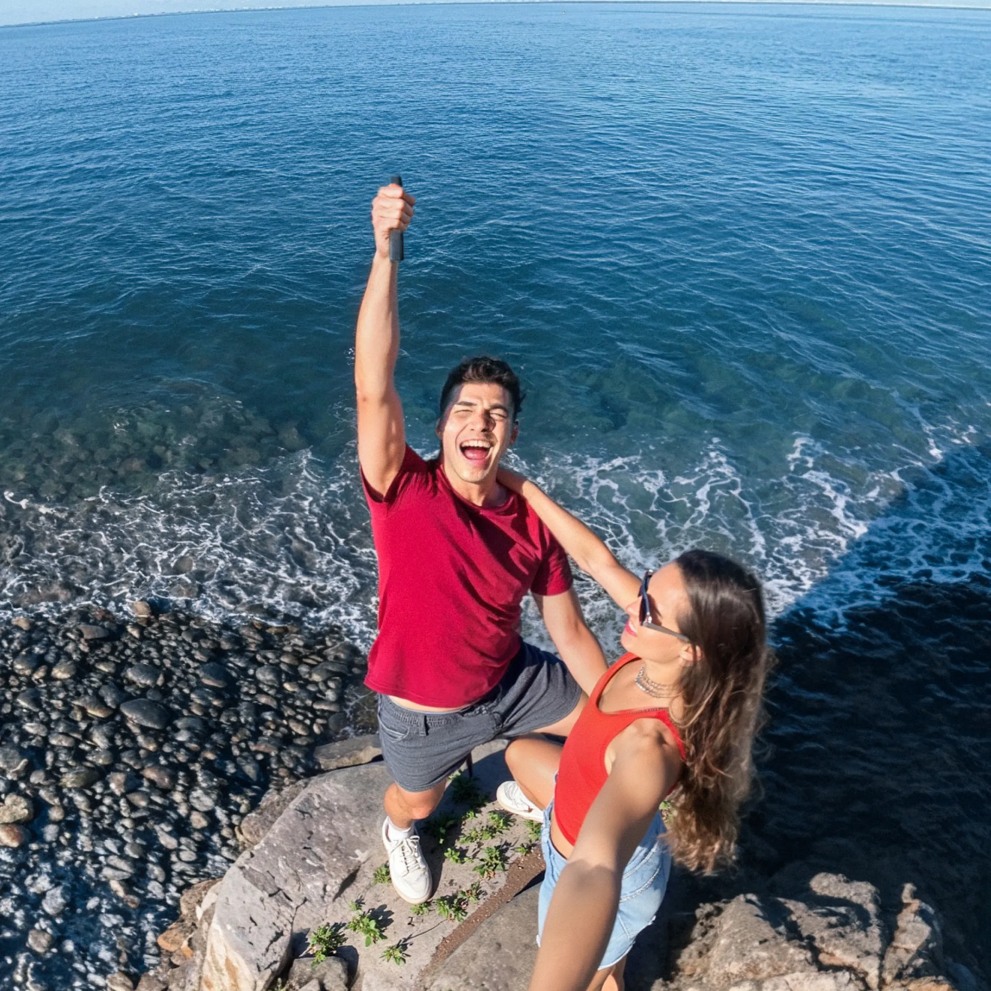 Ein Paar posiert für ein Selfie auf einer felsigen Klippe, der Mann hält ein Telefon hoch und lächelt, mit dem Meer im Hintergrund.