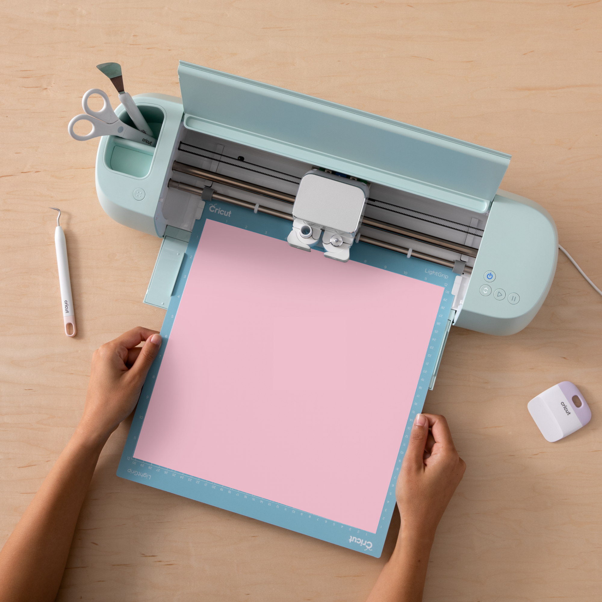 Eine Cricut-Maschine schneidet rosa Papier. Hände halten das Papier auf einer blauen Matte. Werkzeuge und Schere liegen daneben.