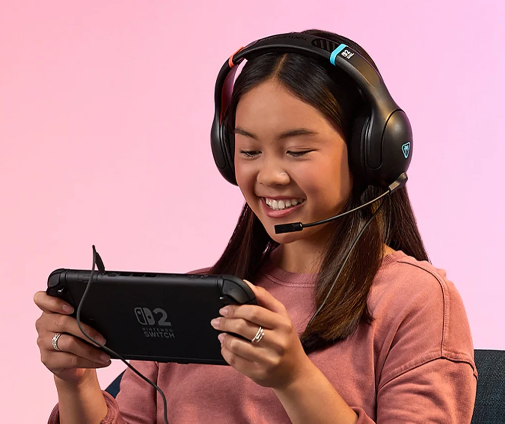 Eine junge Frau lächelt, trägt ein Headset und hält eine Nintendo Switch. Rosa und lila Hintergrund.