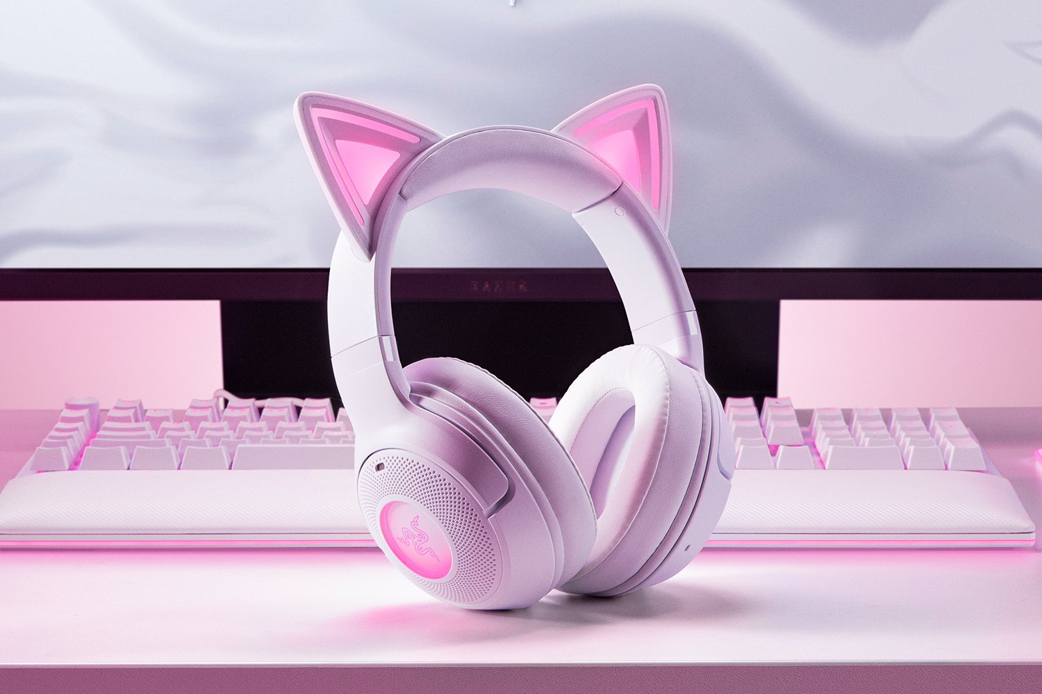 Cuffie bianche con orecchie da gatto su superficie rosa, tastiera e monitor sullo sfondo. Accenti e illuminazione rosa.