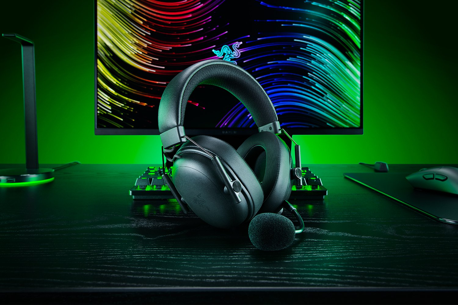Cuffie Razer nere su una scrivania con tastiera, mouse e monitor con linee colorate, il tutto sotto luce verde.
