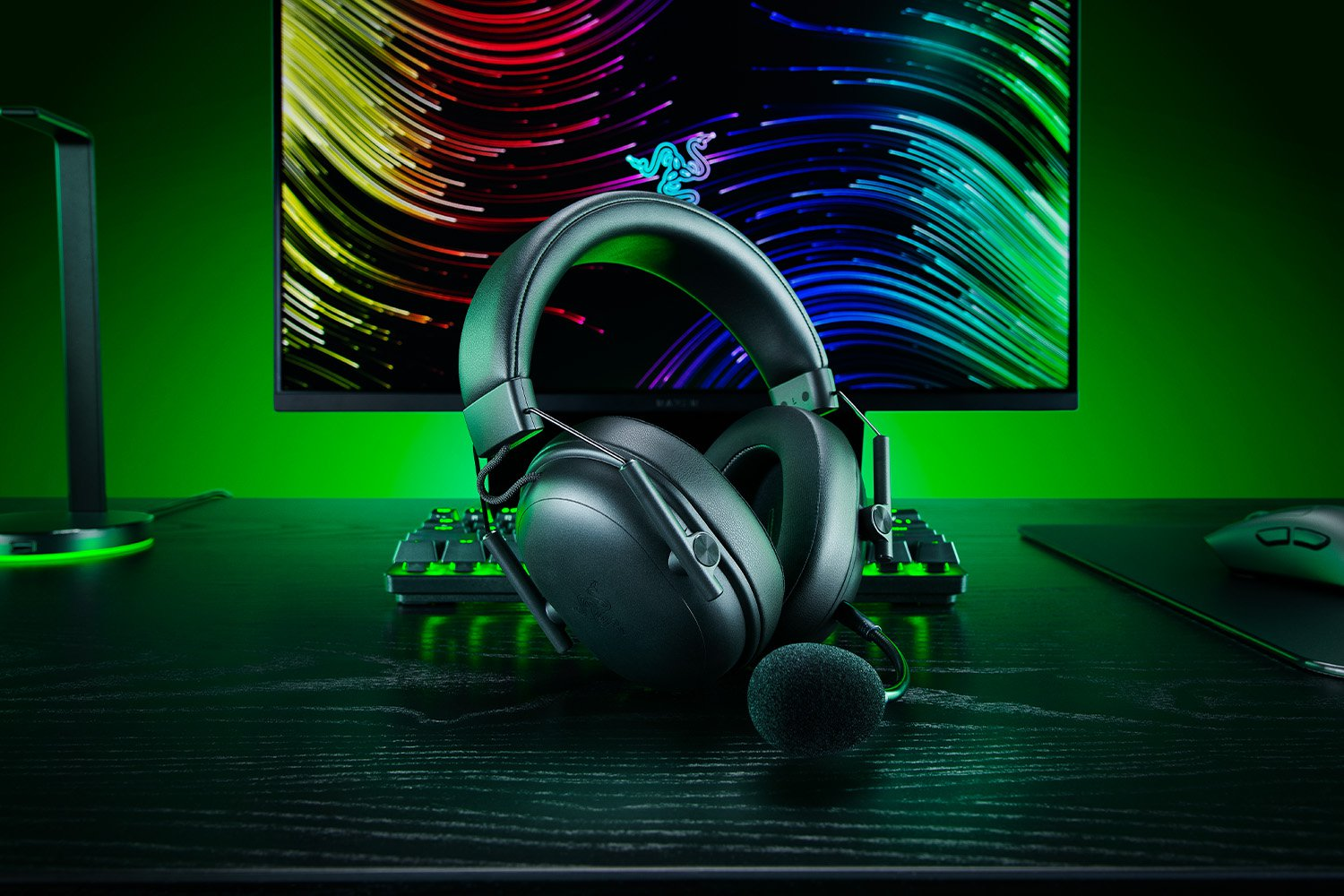 Cuffie da gioco nere con microfono su una scrivania. Una tastiera e un monitor sullo sfondo.