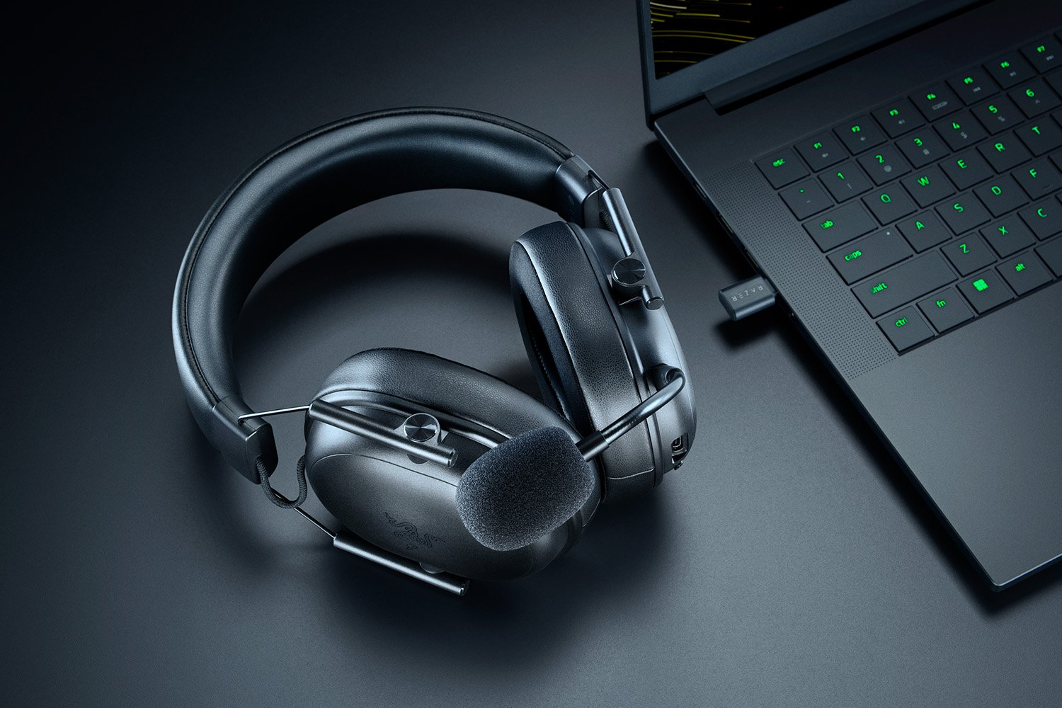 Cuffie da gioco nere con microfono accanto a un laptop. Il laptop è aperto, che mostra una tastiera con tasti retroilluminati verdi.