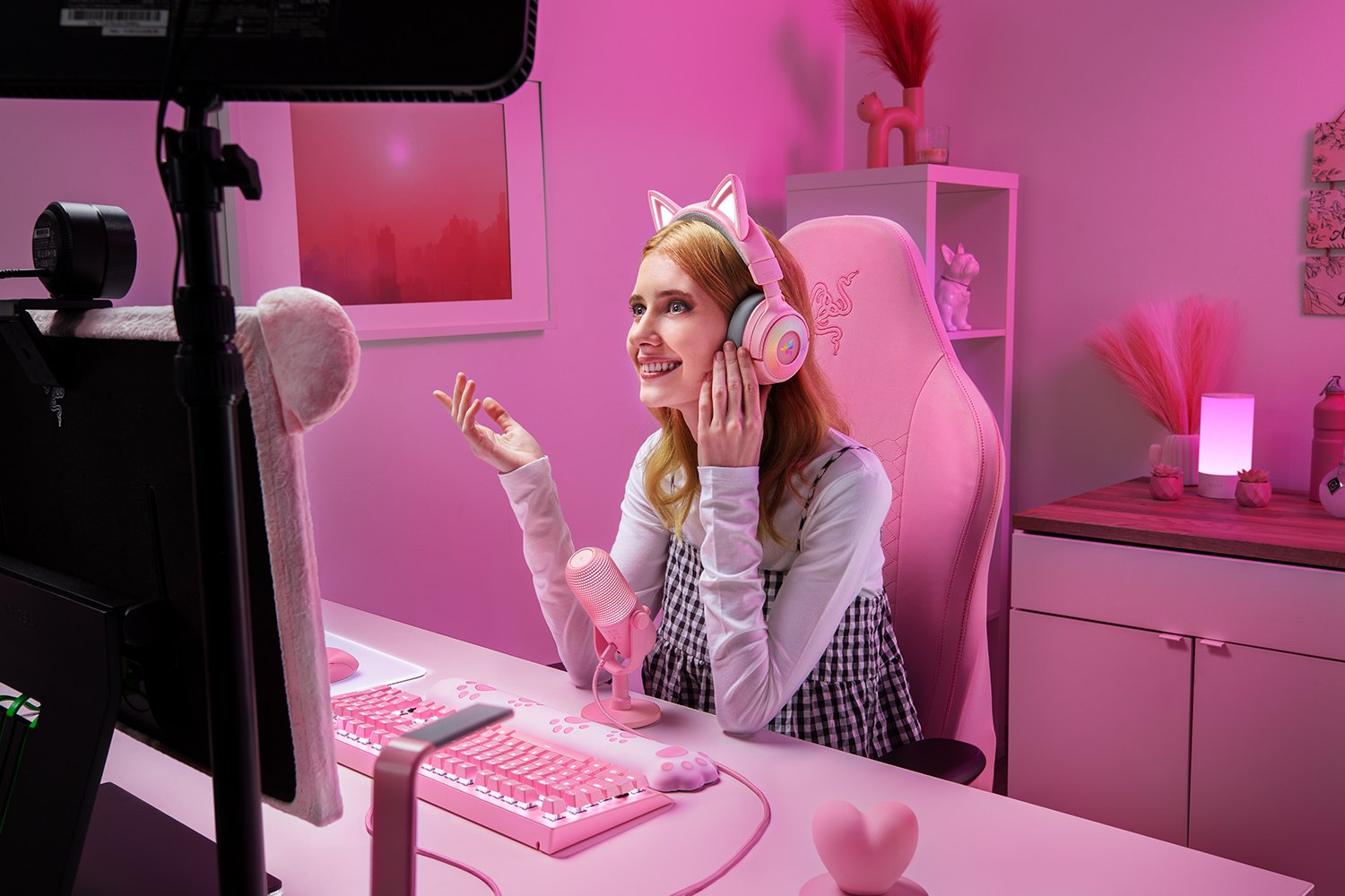Eine Frau mit Katzenohrhörern lächelt. Sie ist in einem rosa Zimmer und benutzt eine rosa Tastatur und ein Mikrofon. Rosa Stuhl und Dekorationen.