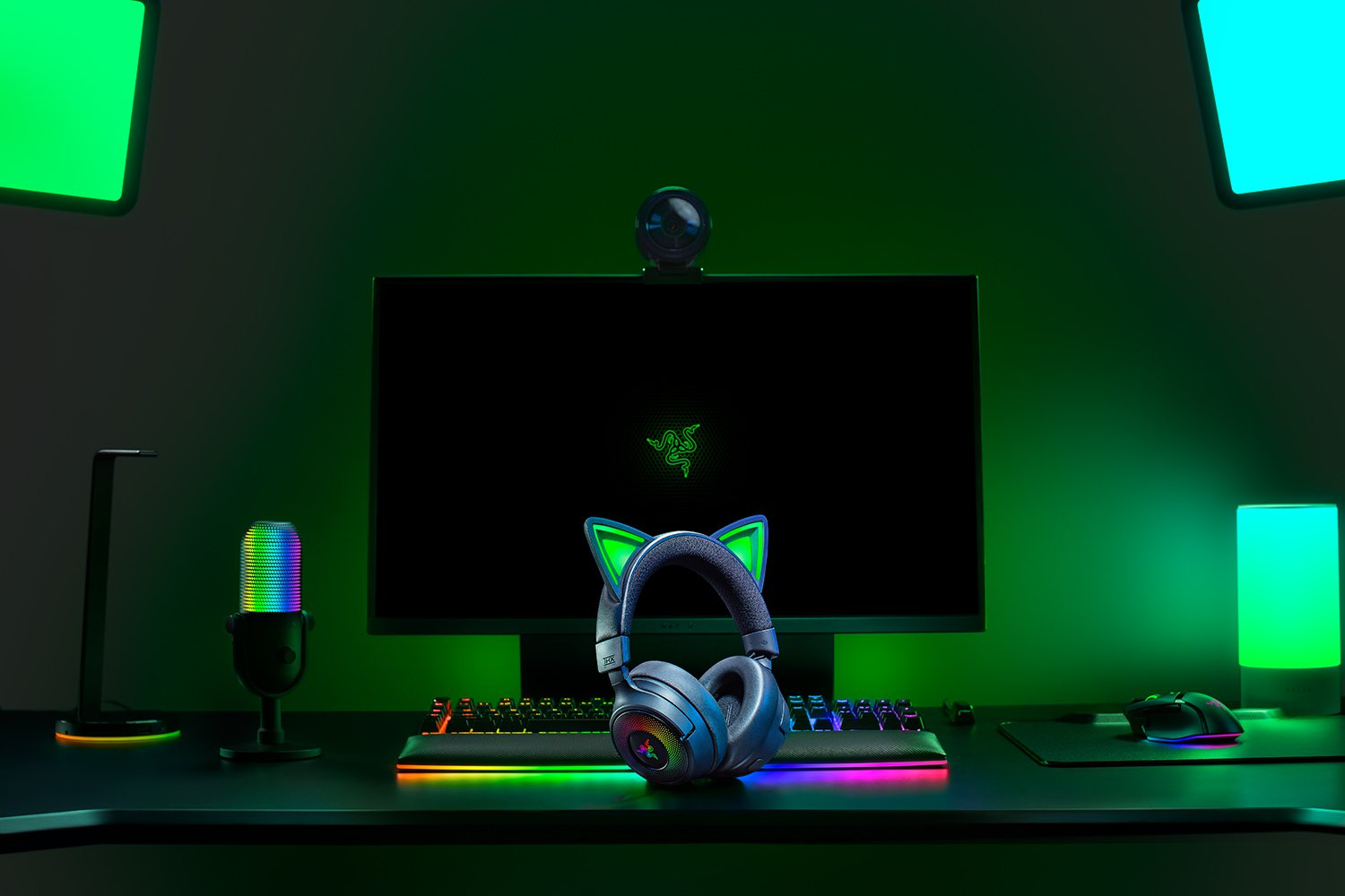 Configurazione di gioco con cuffie, tastiera e monitor Razer. Illuminazione ambientale verde e blu. Sono presenti anche microfono e webcam.