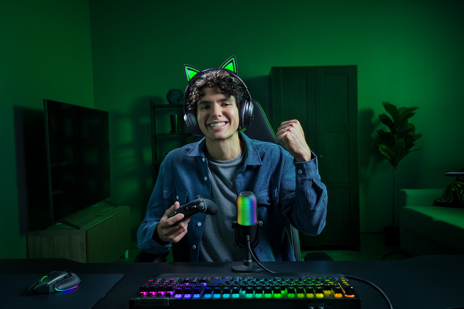 Un giocatore sorridente con cuffie e controller. È in una stanza illuminata di verde, con un microfono e una tastiera.