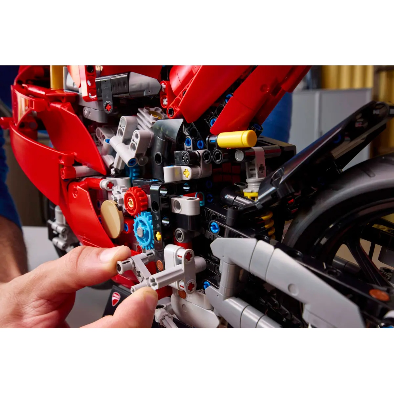 Une personne assemble une moto Lego rouge et noire. Les mains connectent des pièces Lego grises. Des engrenages et autres composants sont visibles.