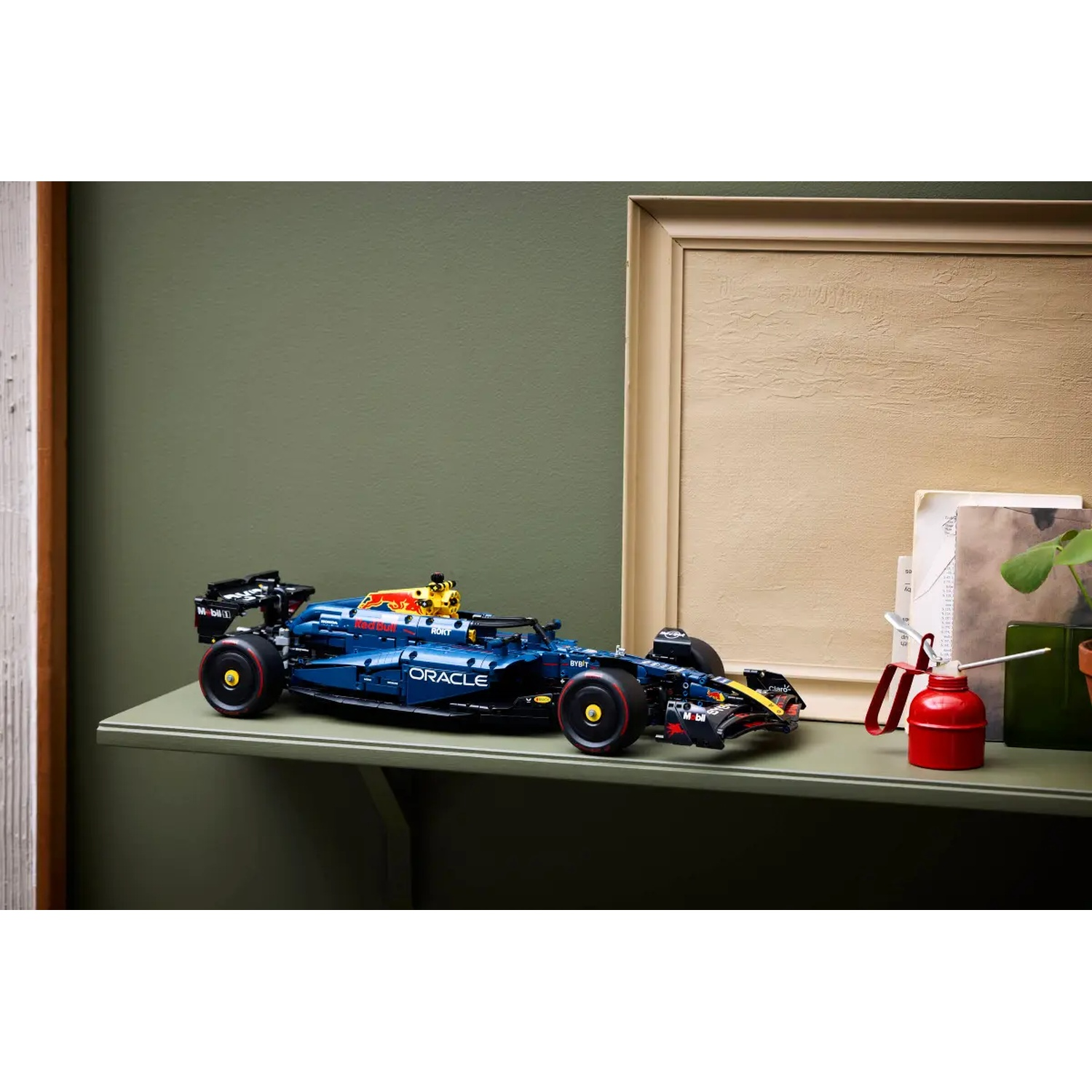 Een donkerblauw Red Bull F1-automodel op een plank met een rode oliekruik en een beige ingelijste foto. Groene muurachtergrond.
