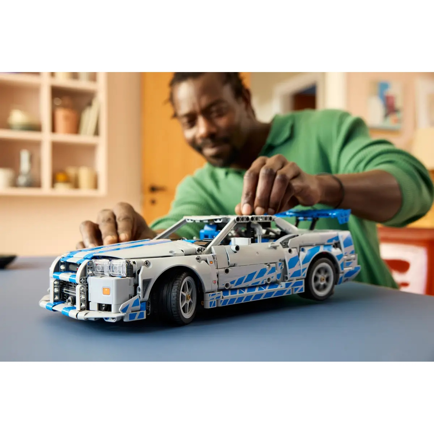 A man assembles a white and blue toy car on a blue table. Background has shelves, a door and a painting. He is wearing green.