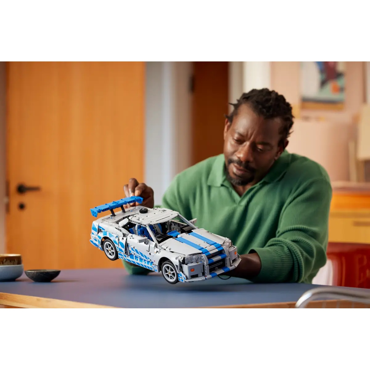 A man holds a white and blue toy car. He wears a green shirt. The car is on a blue table. Background is blurred.