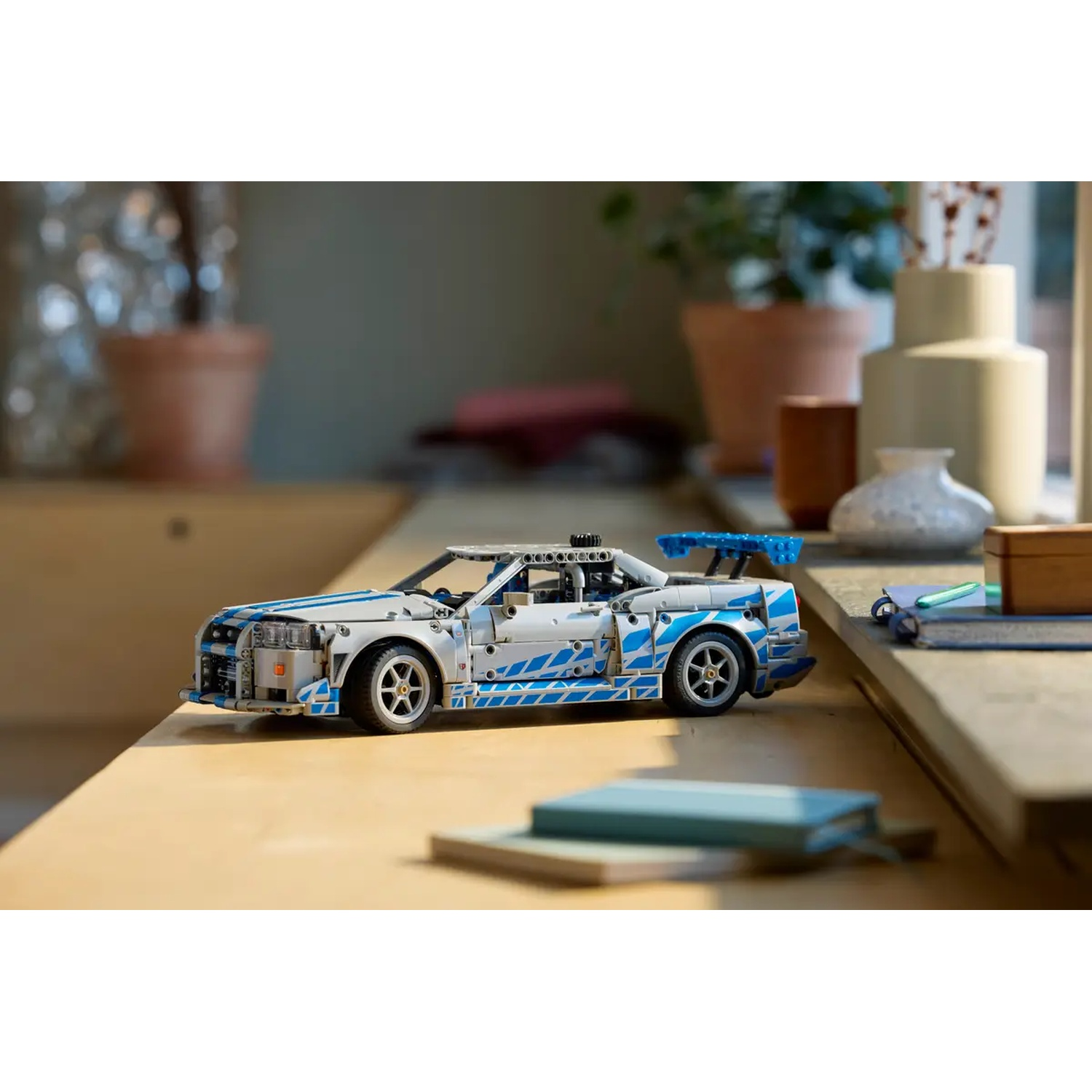 A white and blue toy car is on a wooden surface. Plants and vases are in the background, near a window.