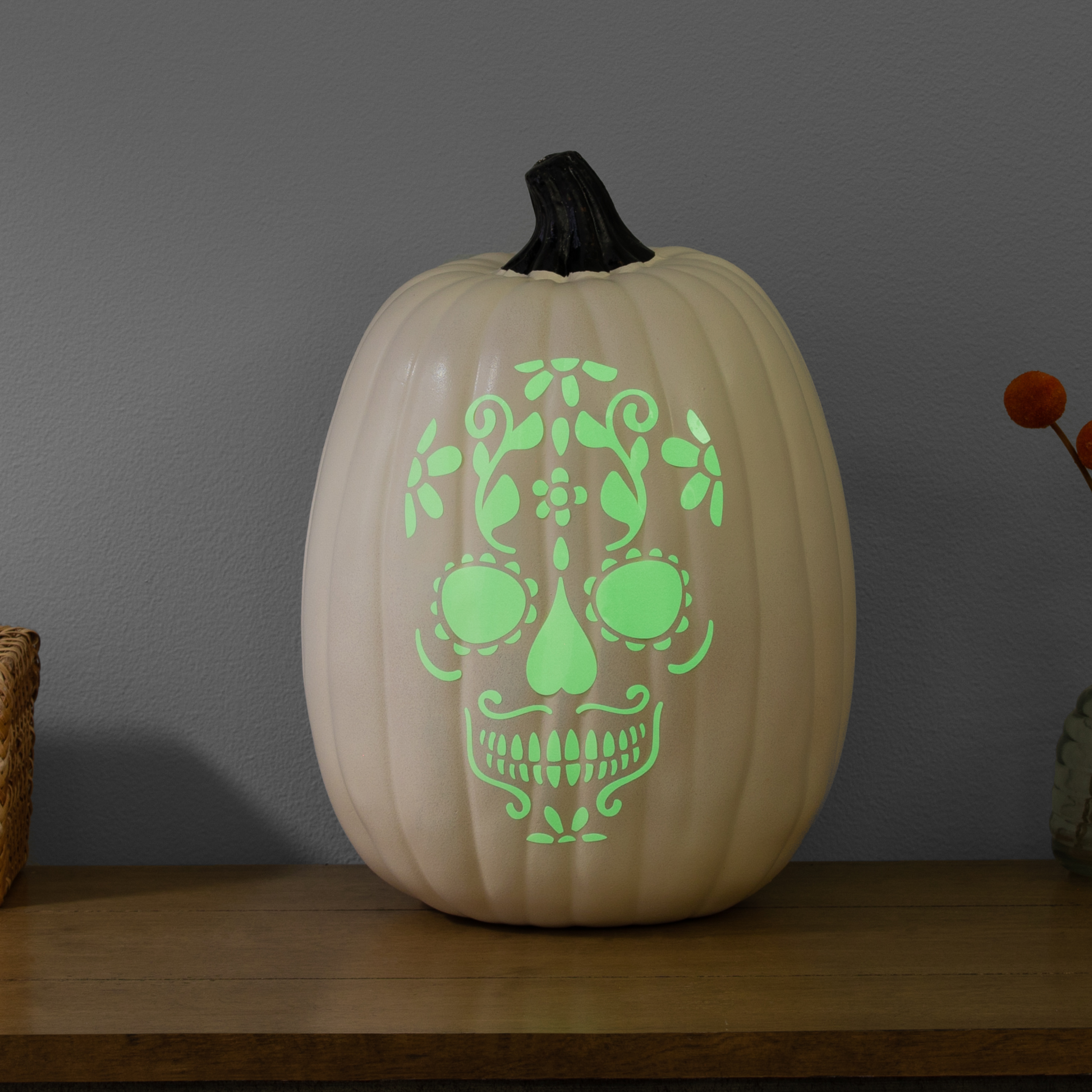 Ein beiger Kürbis mit einem leuchtend grünen Totenkopf-Design. Auf einer Holzoberfläche vor einer grauen Wand.