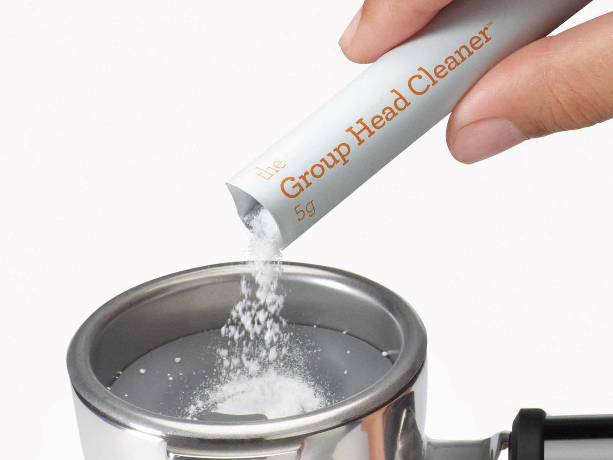 Egy kéz tisztítószert önt egy tasakból egy eszpresszó gép szűrőtartójába. A tasakon a 'Group Head Cleaner' felirat látható.