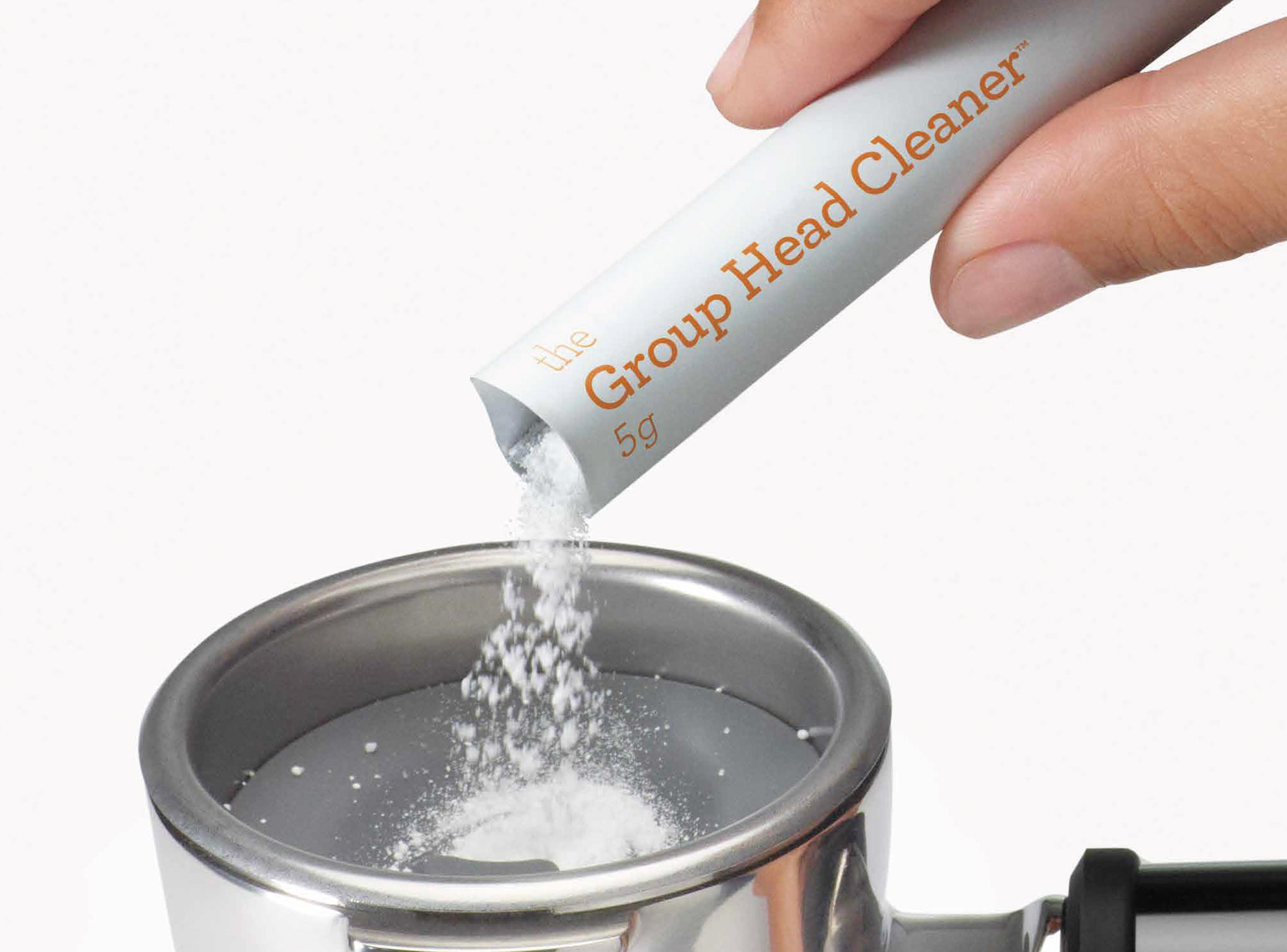 Egy kéz tisztítószert önt a 'Group Head Cleaner' feliratú tasakból egy kávéfőző szűrőtartójába.