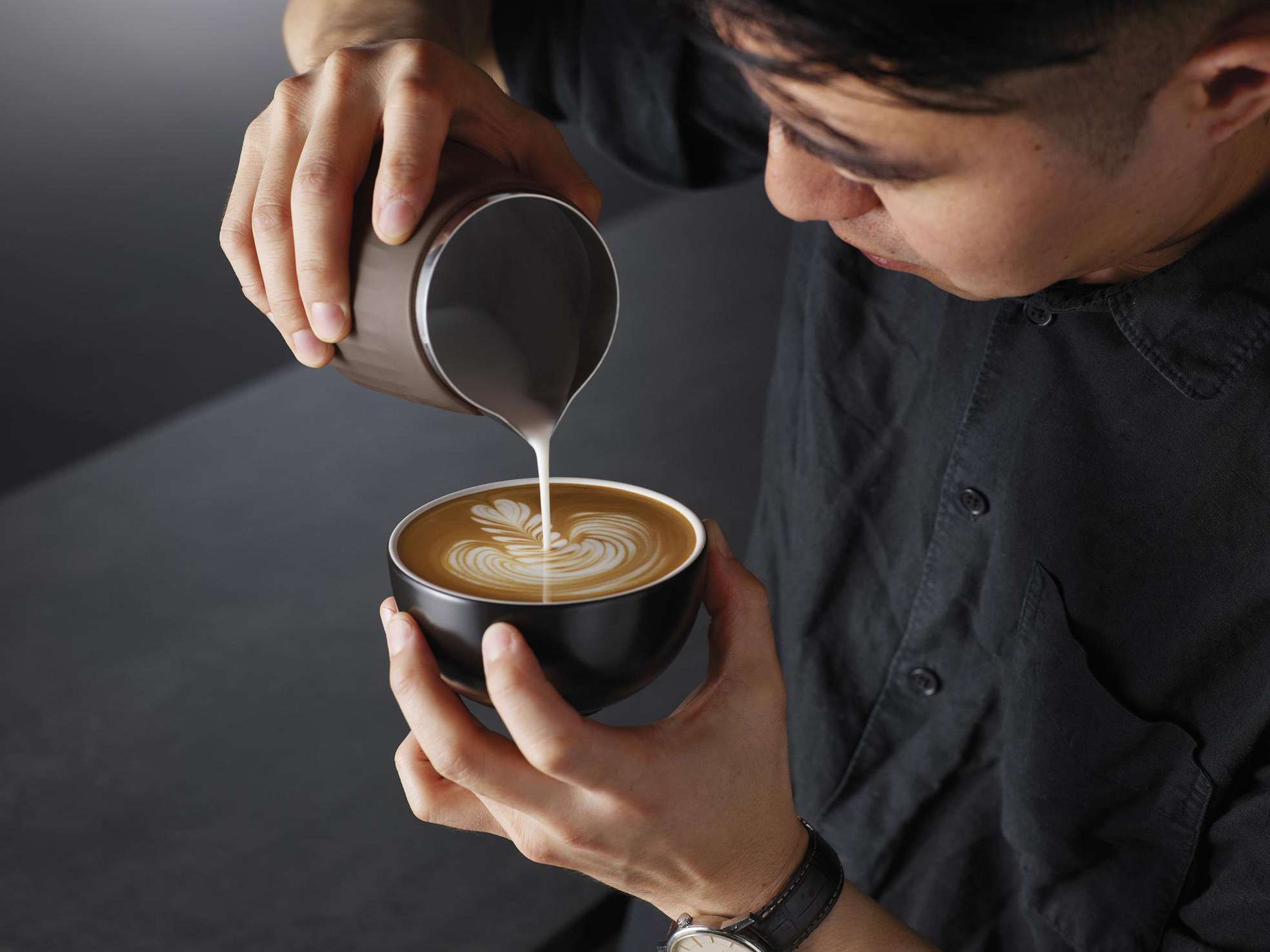 Egy barista tejet önt kávéscsészébe, latte artot készítve. A csészét a barista tartja a kezében, sötét háttérrel.