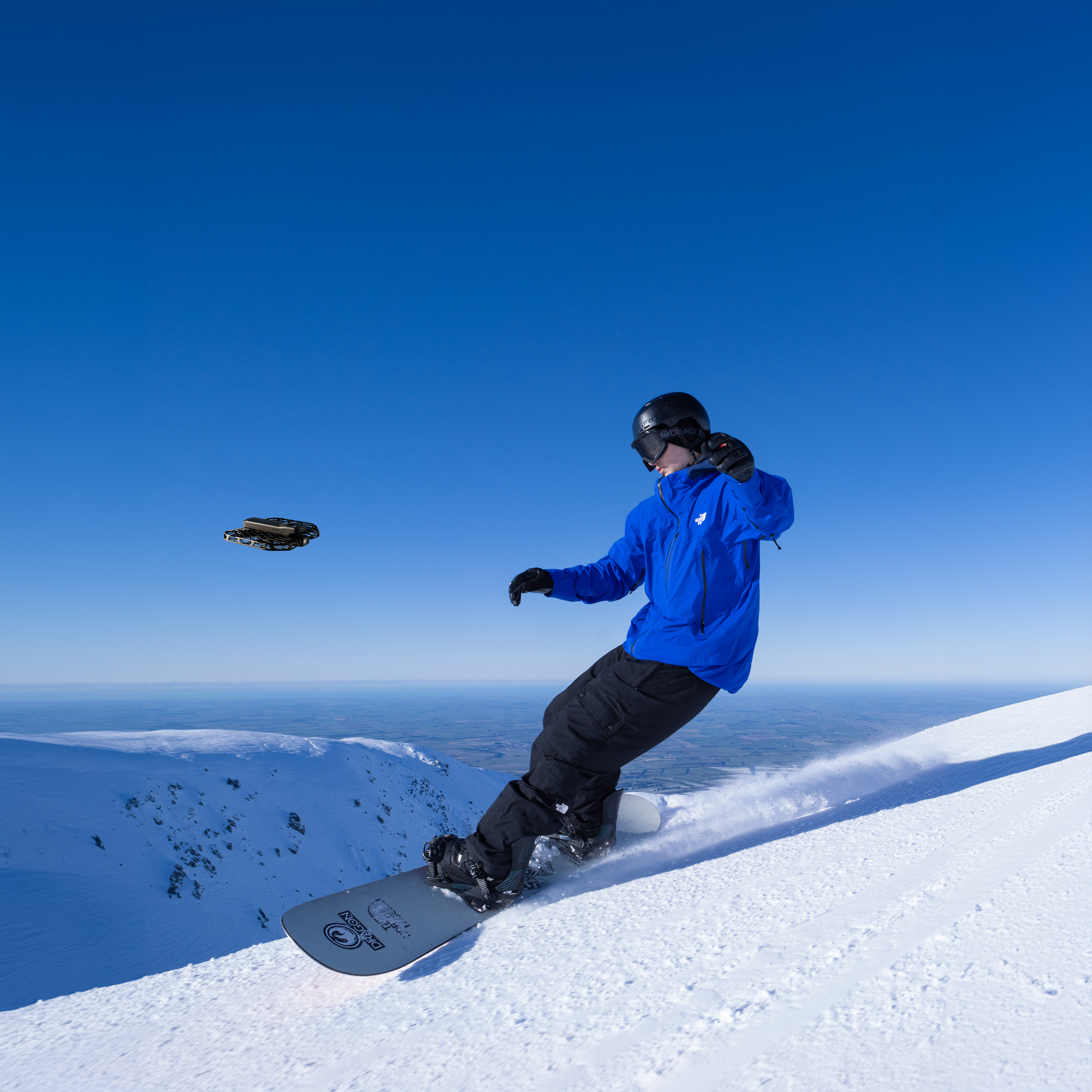 Ein Snowboarder in blauer Jacke fährt unter blauem Himmel einen schneebedeckten Hang hinunter. Eine Drohne ist am Himmel.