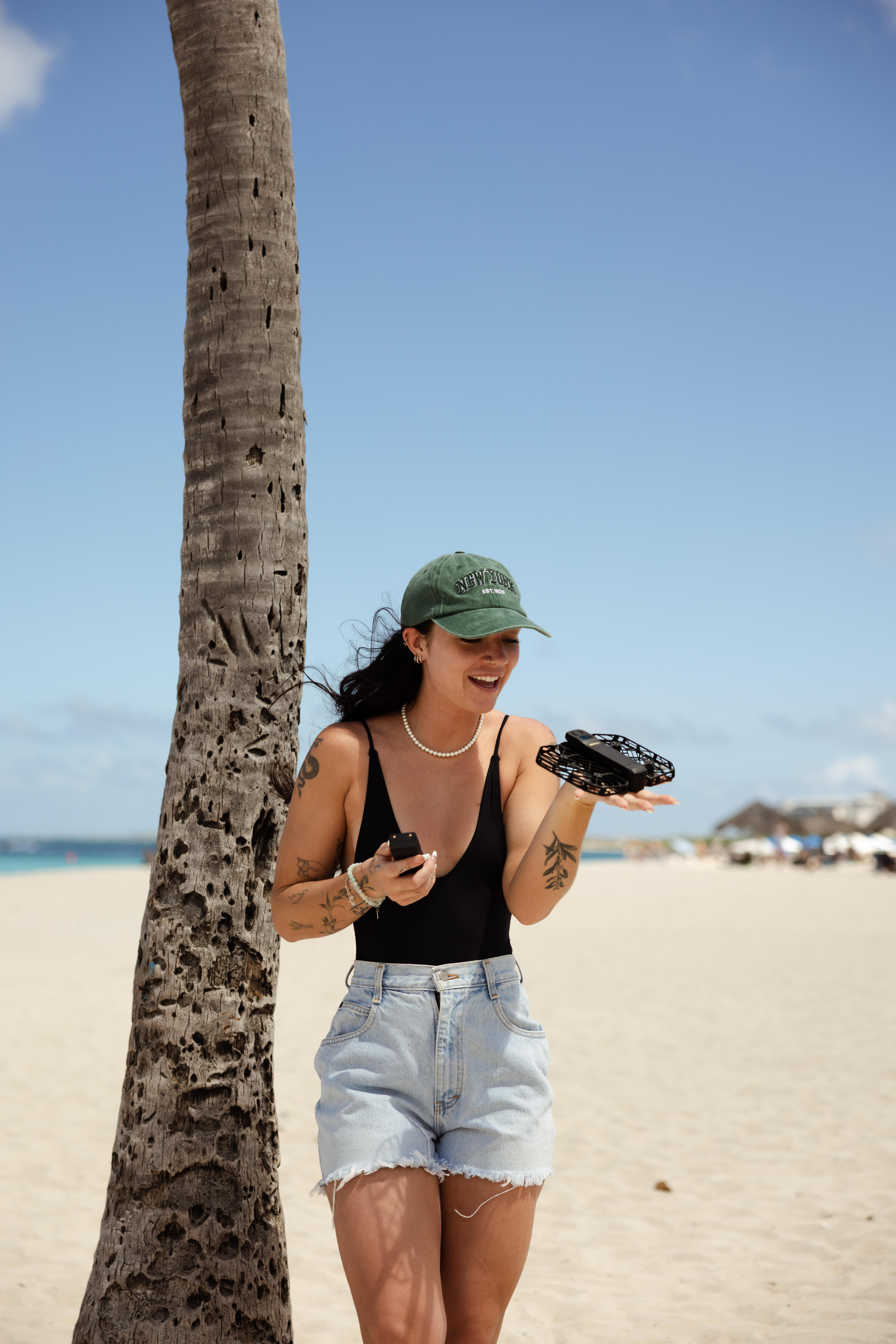 Frau am Strand, die eine Drohne und ein Telefon hält. Sie trägt eine Kappe und Jeansshorts, neben ihr eine Palme.