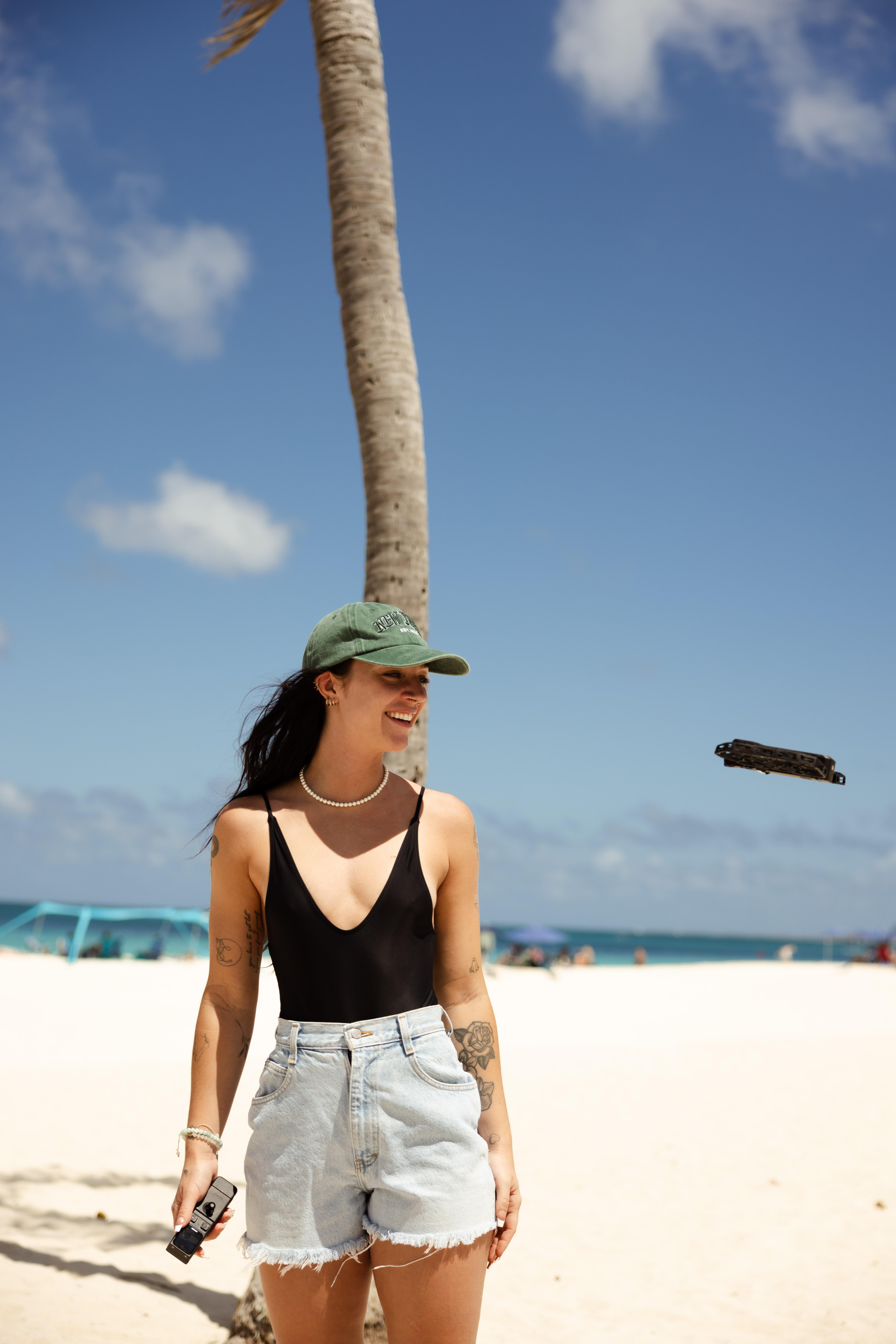 Frau in schwarzem Badeanzug, Jeansshorts und grüner Kappe lächelt am Strand. Palme und blauer Himmel im Hintergrund.