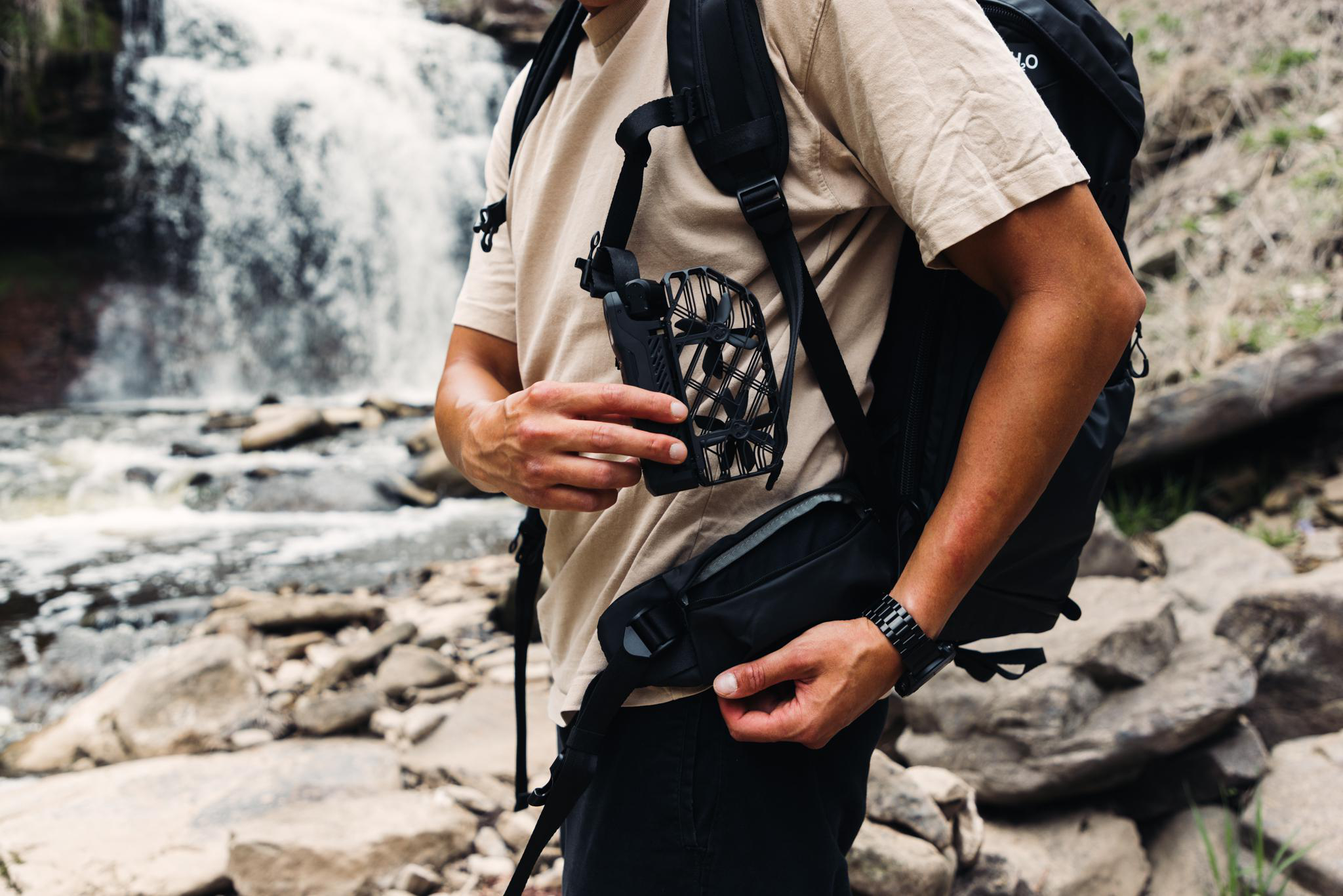 Eine Person hält ein schwarzes Gerät mit einem Rucksack in der Nähe eines Wasserfalls. Die Person trägt ein hellbraunes Hemd und eine schwarze Hose.