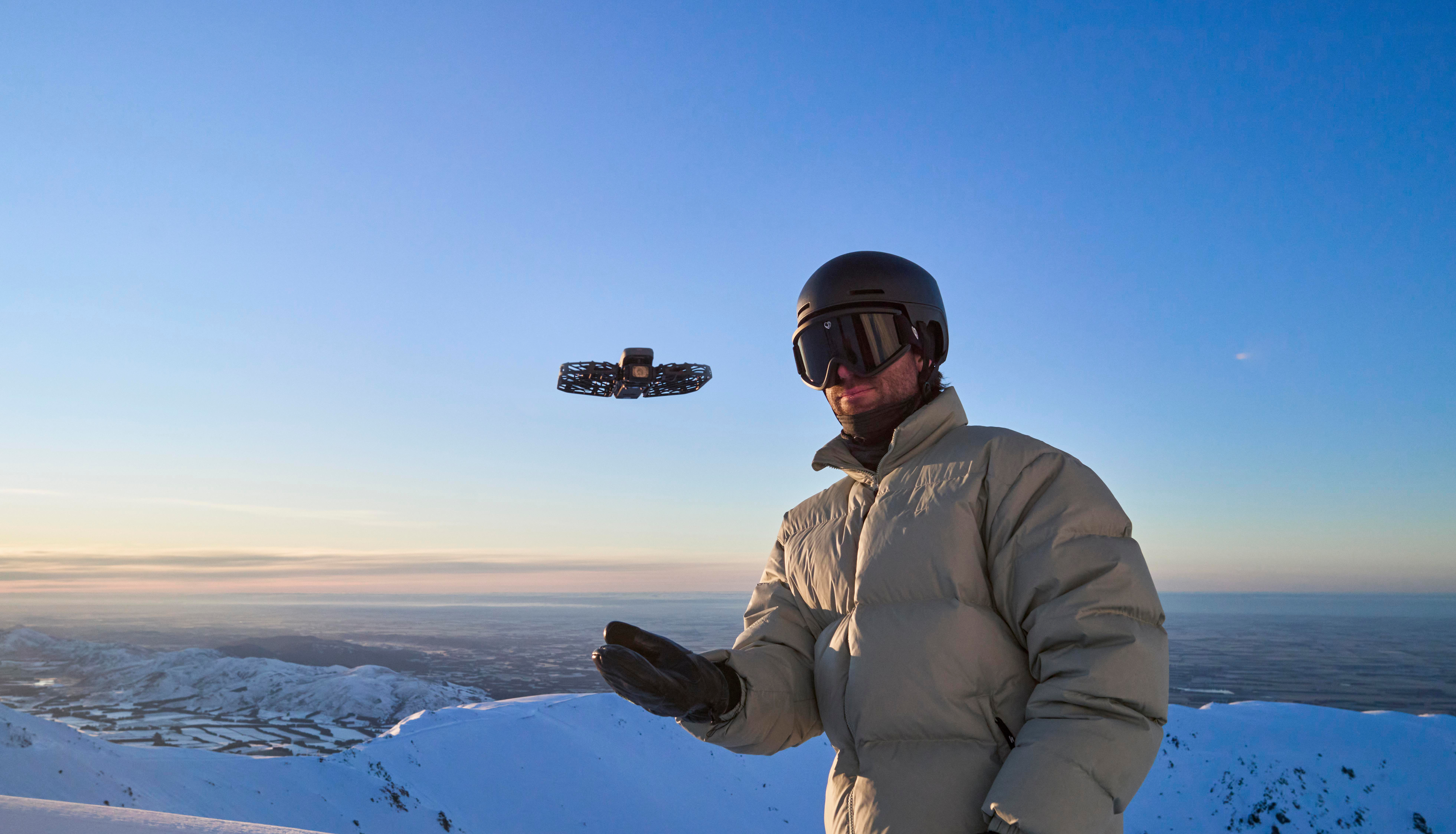 Mann in Skiausrüstung mit Drohne über schneebedeckten Bergen. Drohne in der Luft. Klarer Himmel. Der Mann trägt Helm und Brille.