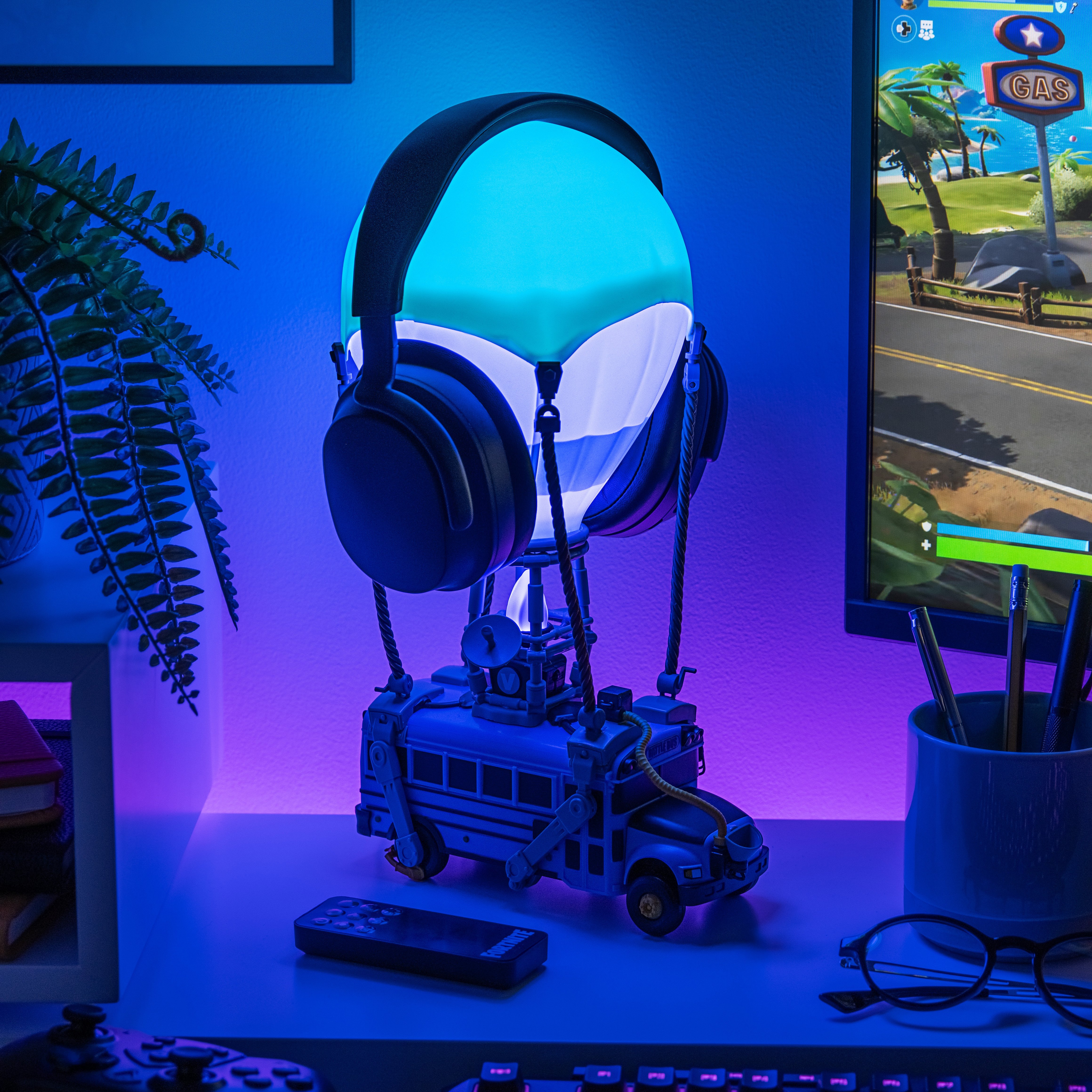 Eine Fortnite-Lampe mit Kopfhörern, ein Spielbildschirm und ein Farn auf einem Schreibtisch mit Controller und Tastatur.