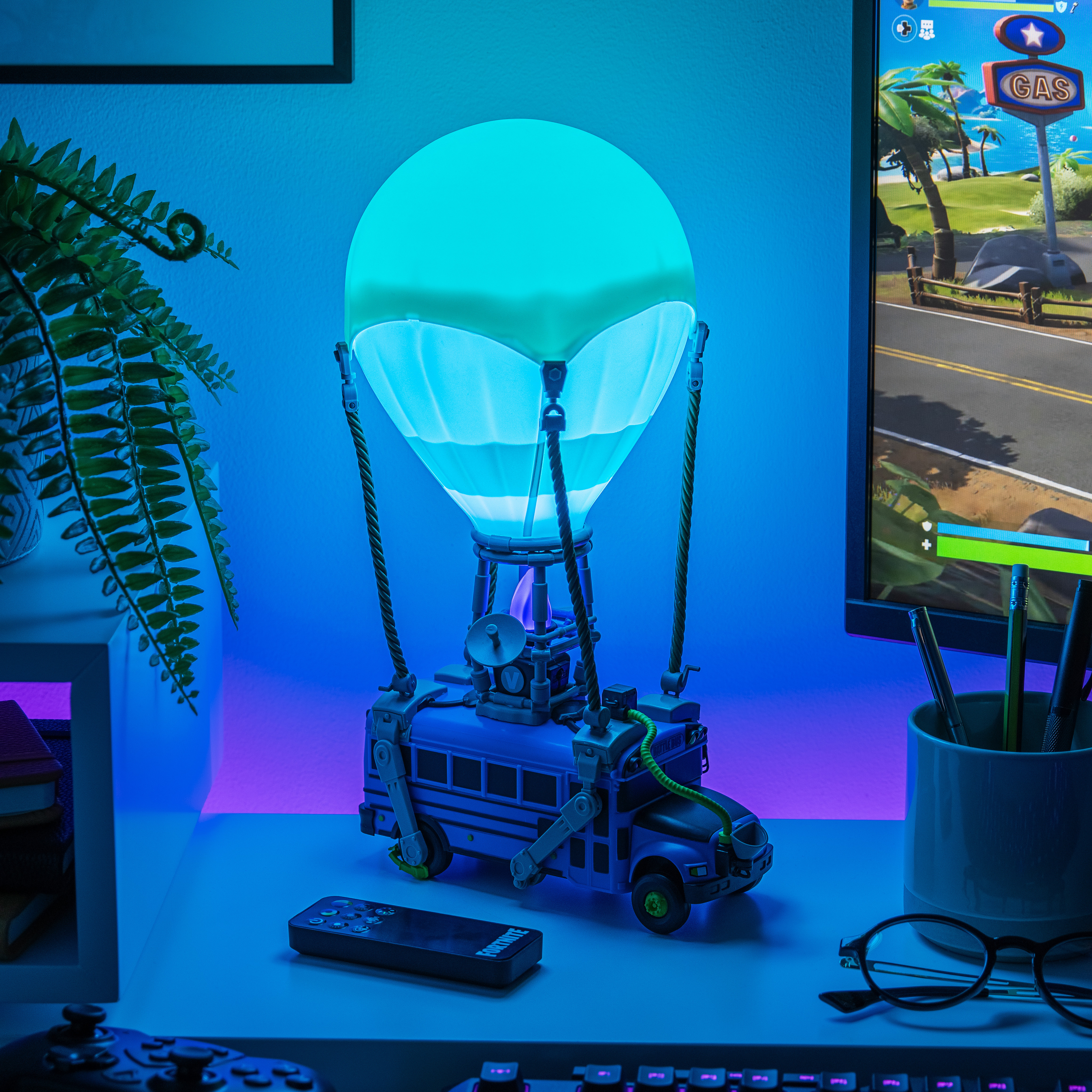Eine Fortnite Battle Bus Lampe leuchtet auf einem Schreibtisch. Ein Controller, eine Fernbedienung und ein Farn sind ebenfalls sichtbar.
