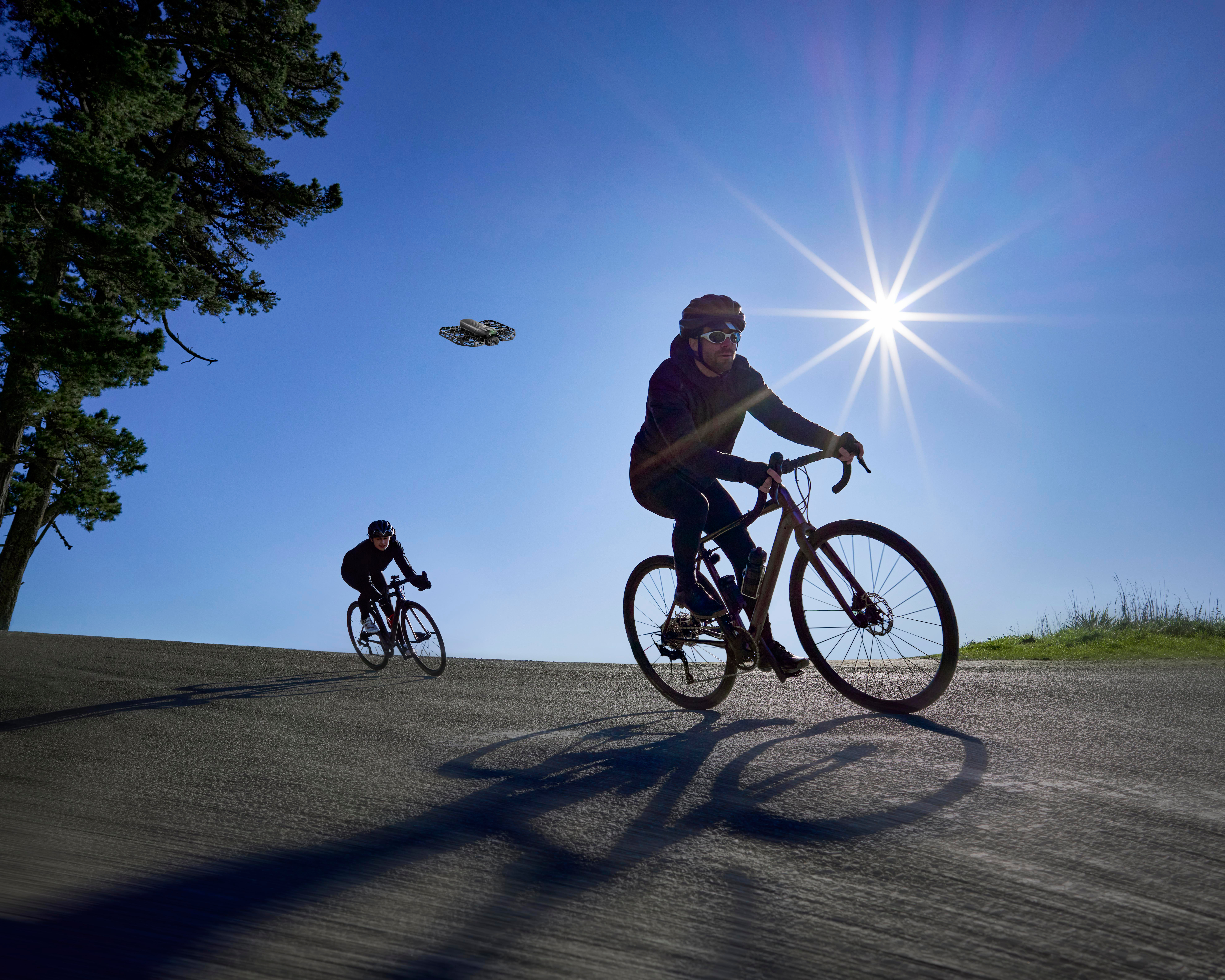 Zwei Radfahrer fahren auf einer Straße, im Gegenlicht vor einer hellen Sonne am blauen Himmel. Links ein Baum, im Himmel eine Drohne.