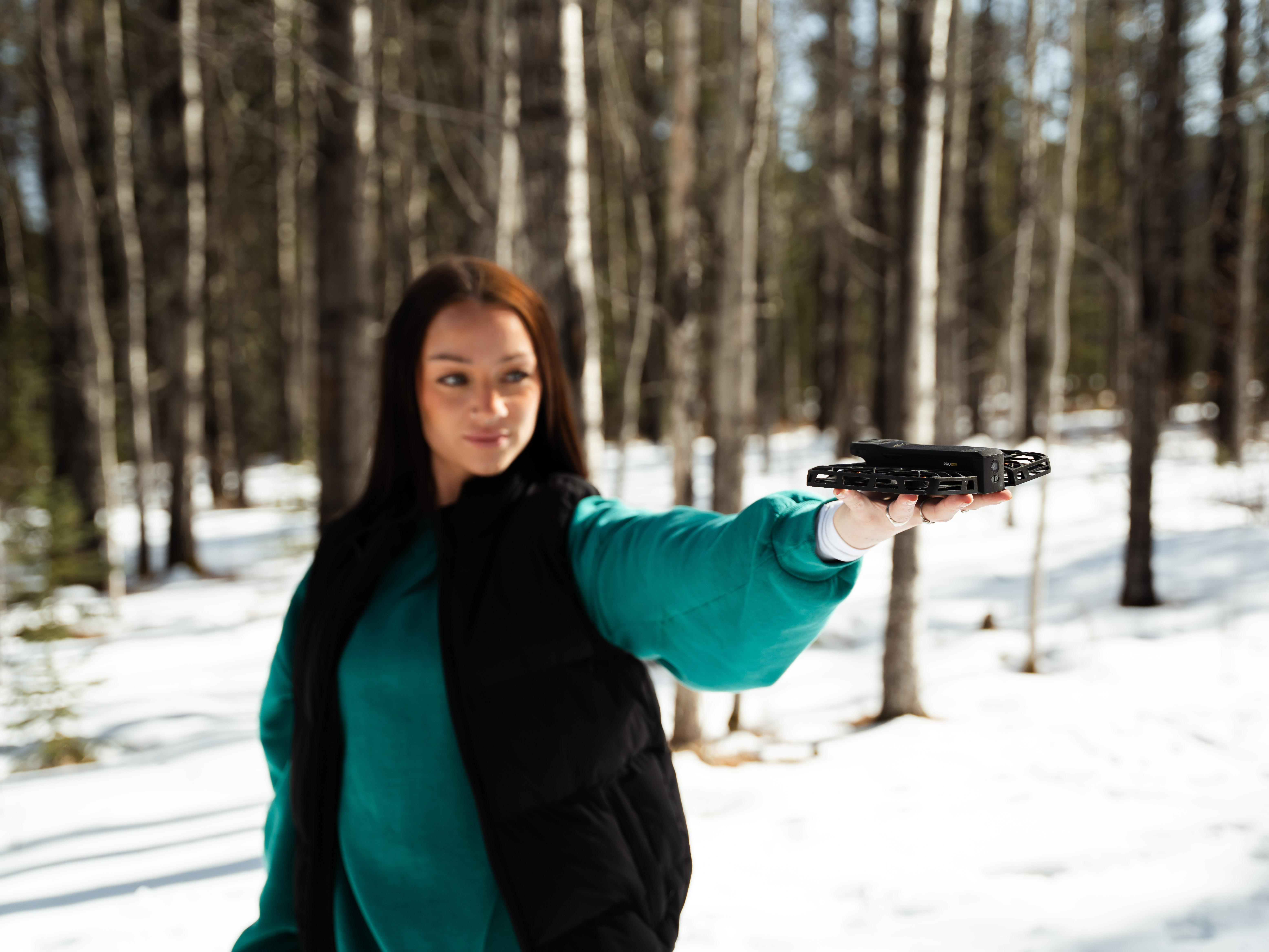 Eine Frau in Türkis und Schwarz hält eine schwarze Drohne in einem verschneiten Wald. Bäume sind im Hintergrund.