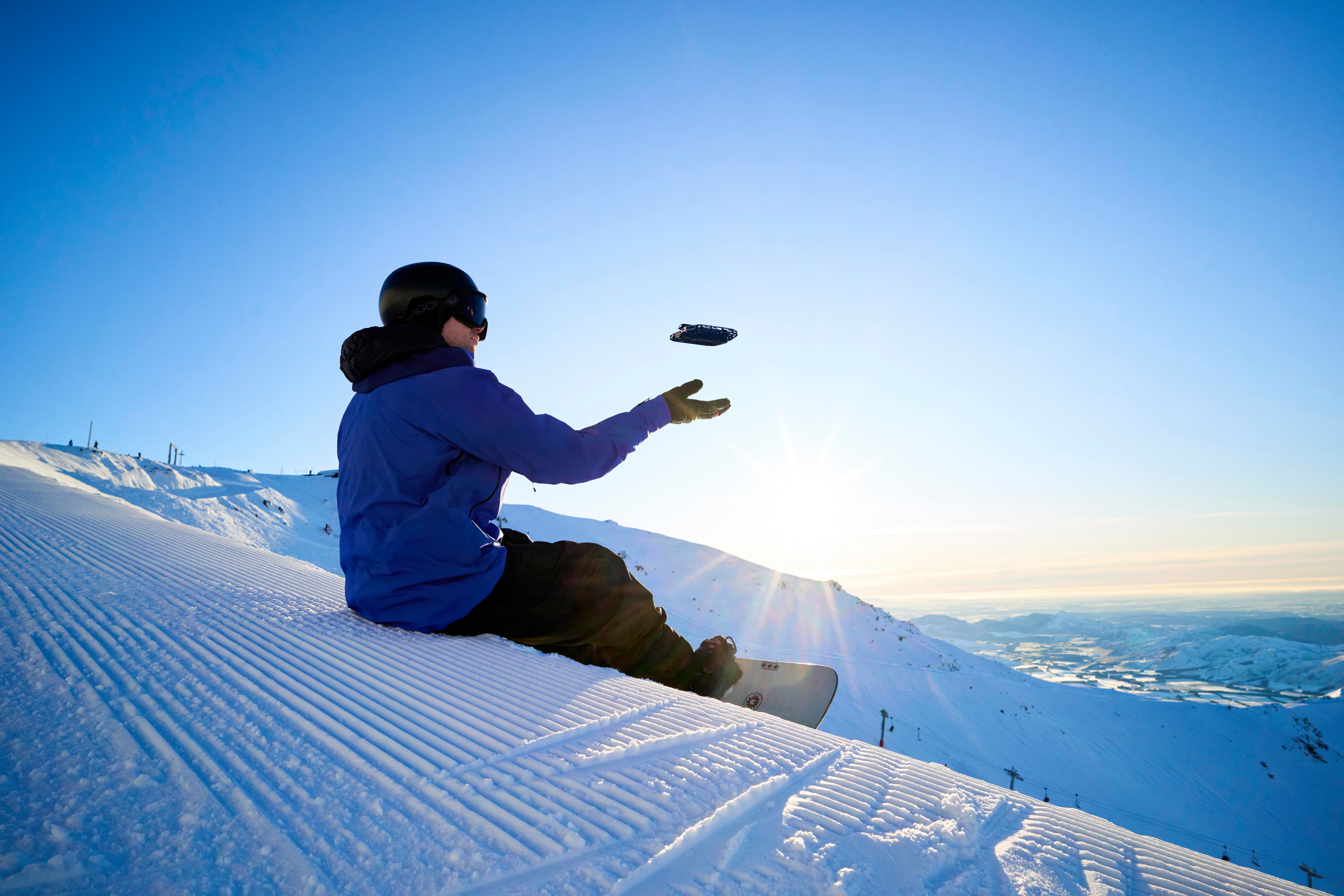 Ein Snowboarder sitzt auf einem schneebedeckten Hang und greift nach einer Drohne vor einem strahlend blauen Himmel, Berge im Hintergrund.