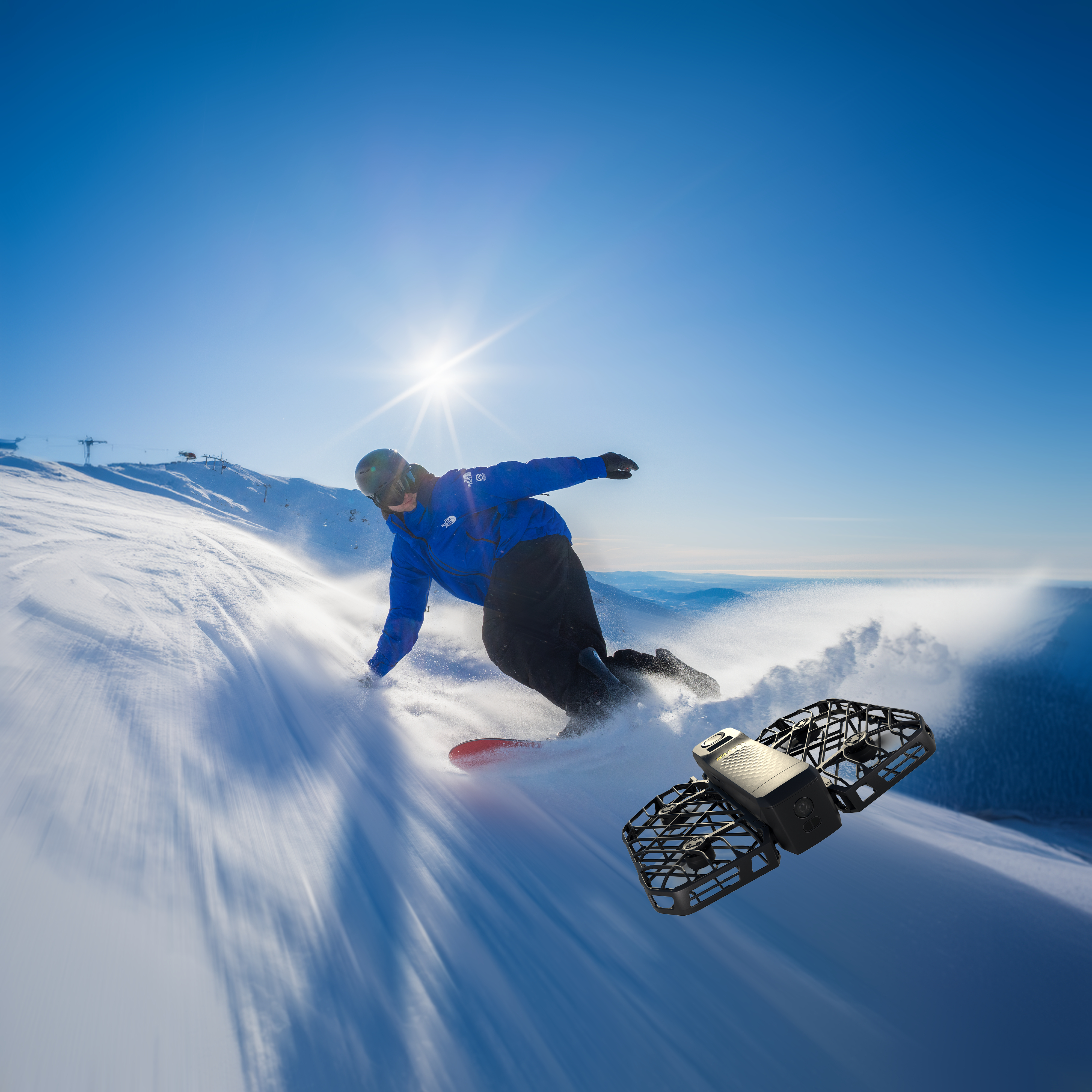 Snowboarder in blauer Jacke fährt einen verschneiten Hang hinunter. Eine Drohne schwebt in der Nähe.
