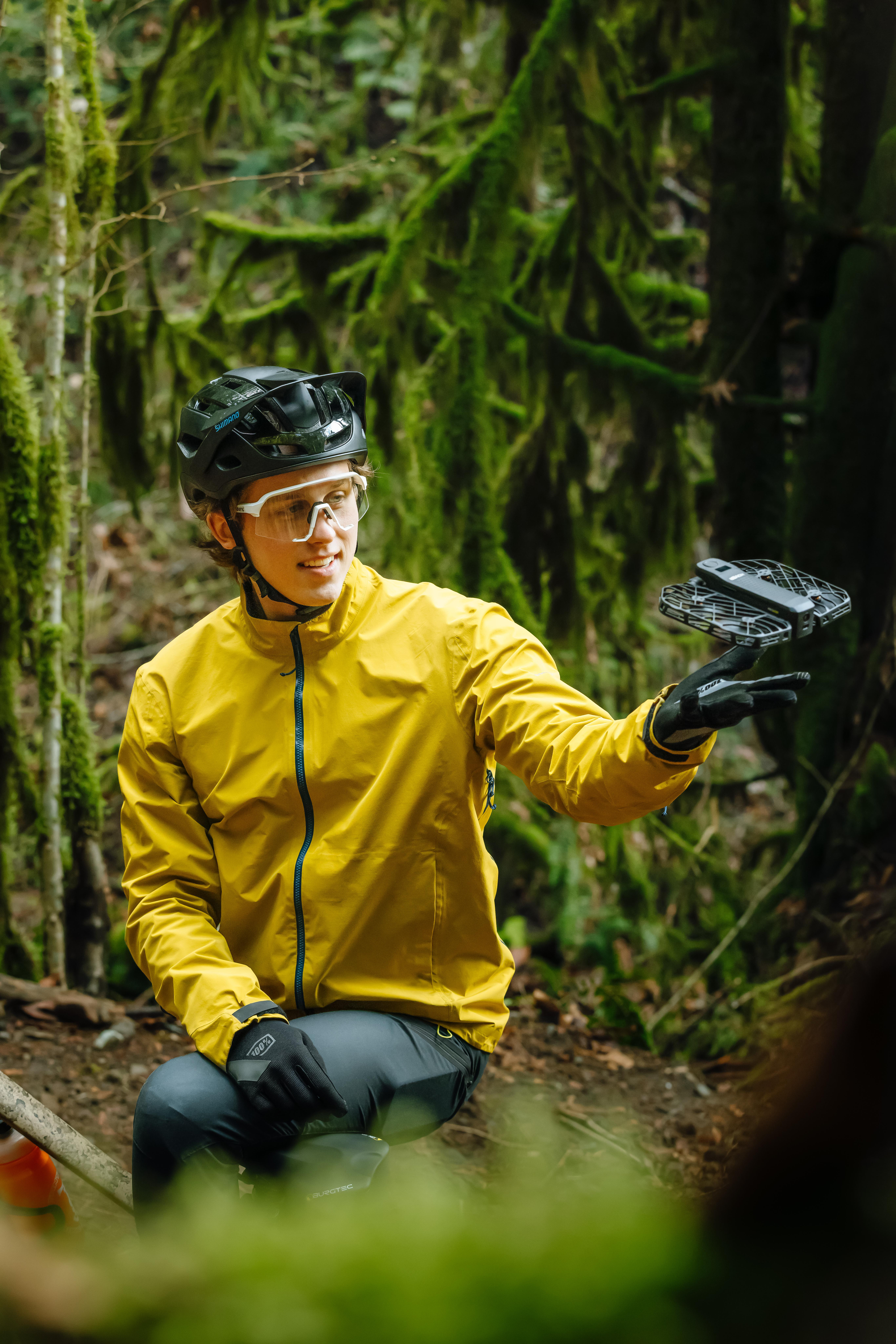 Eine Person in gelber Jacke, schwarzem Helm und Handschuhen hält eine kleine Drohne in einem Wald mit moosbedeckten Bäumen.