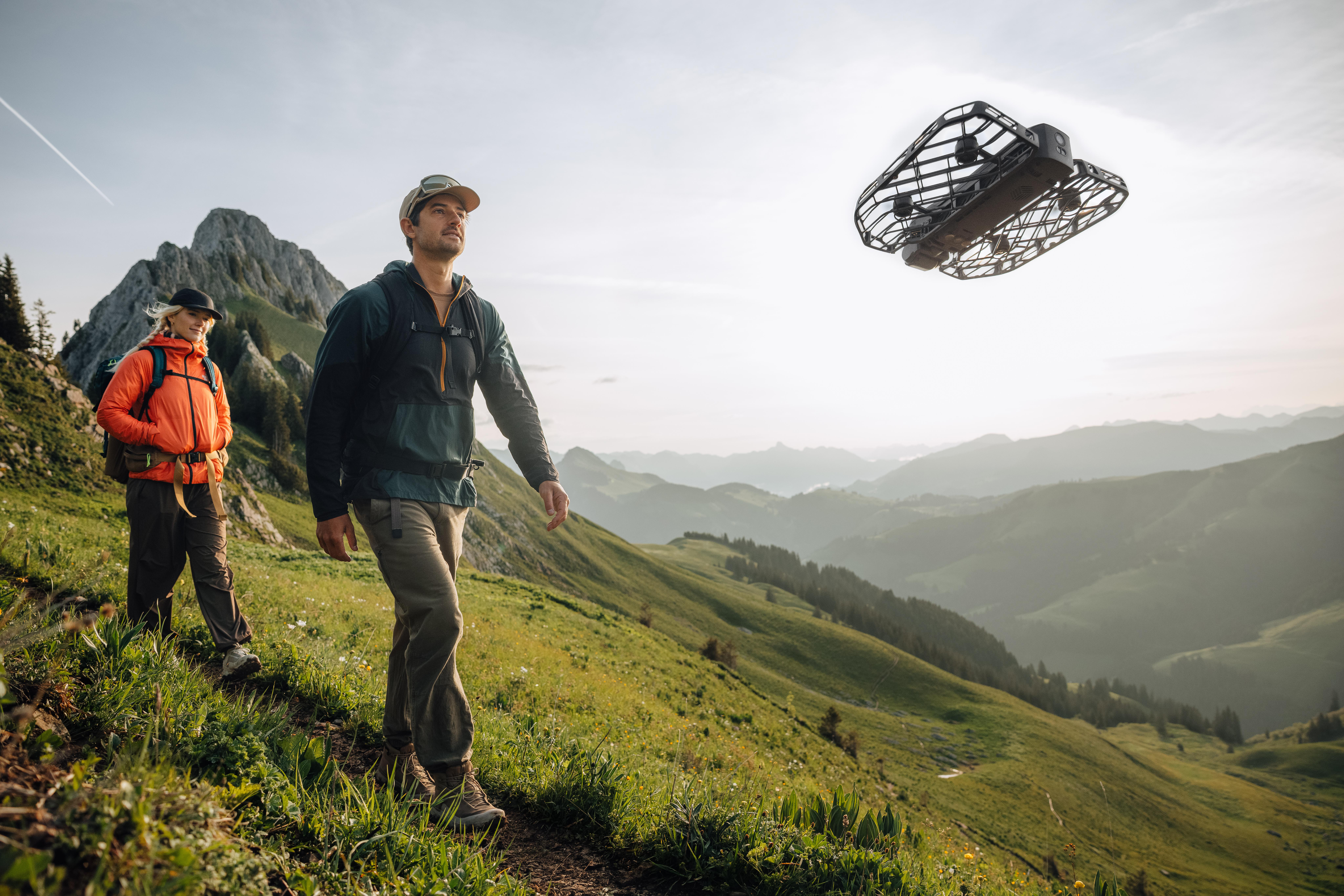 Zwei Wanderer auf einem grünen Hügel, Drohne am Himmel. Einer trägt Orange, der andere Grün. Berge im Hintergrund.