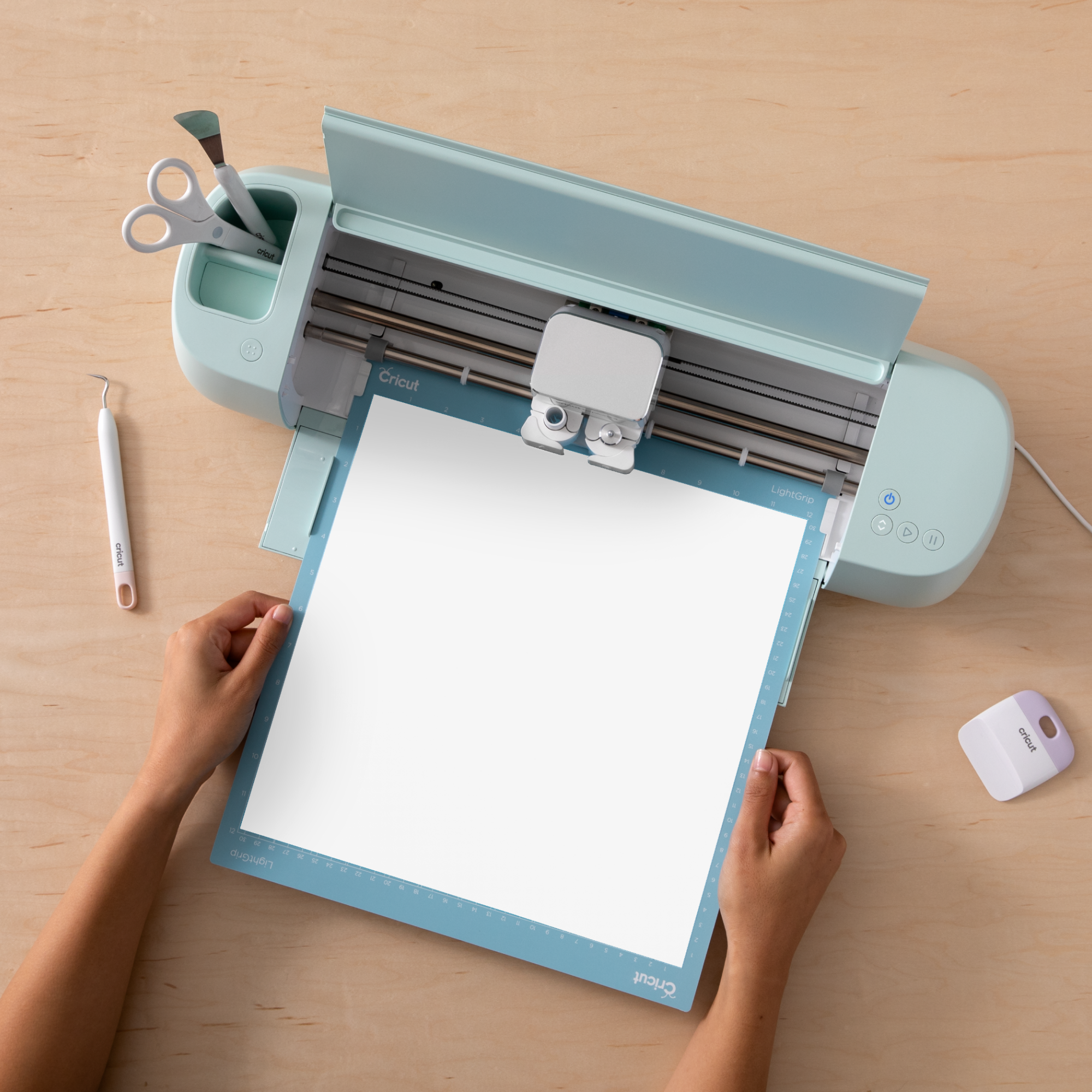 Eine Cricut-Maschine mit Schneidematte und Papier. Hände halten die Matte. Werkzeuge und Zubehör sind in der Nähe.
