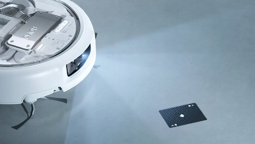 Biały odkurzacz automatyczny obok karty Asa Pik na szarej powierzchni.