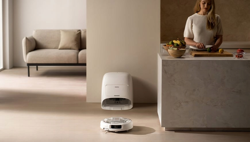 Kobieta kroi warzywa w kuchni. Robot sprzątający jest na podłodze z stacją ładującą. W tle kanapa.