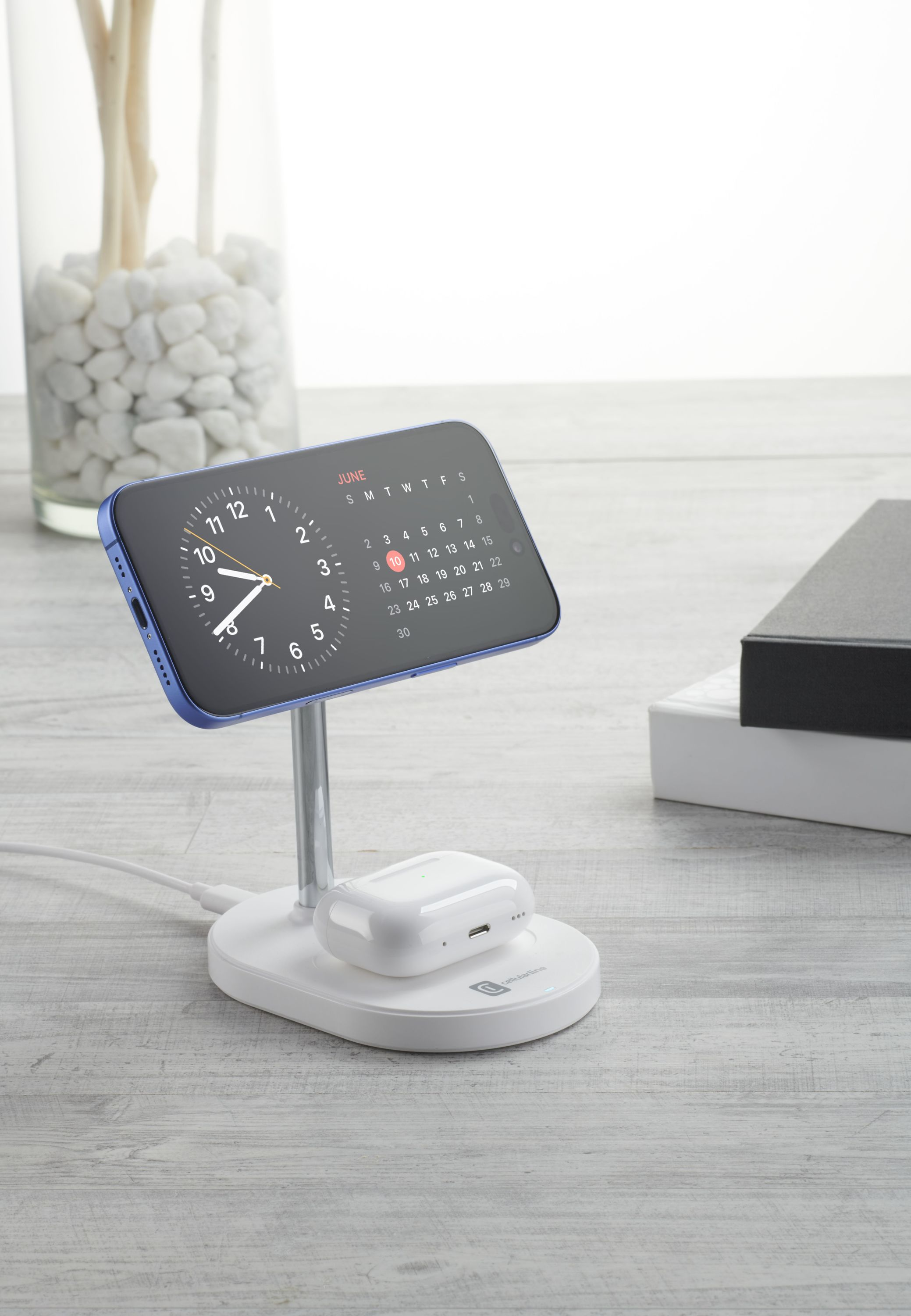 Telefono e auricolari su un caricatore, con orologio e calendario di giugno. Un vaso con pietre bianche sullo sfondo.