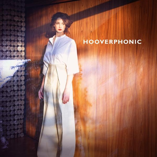 Eine Frau in weißem Hemd und Hose steht vor einer Holzwand. Der Text 'HOOVERPHONIC' ist rechts sichtbar.