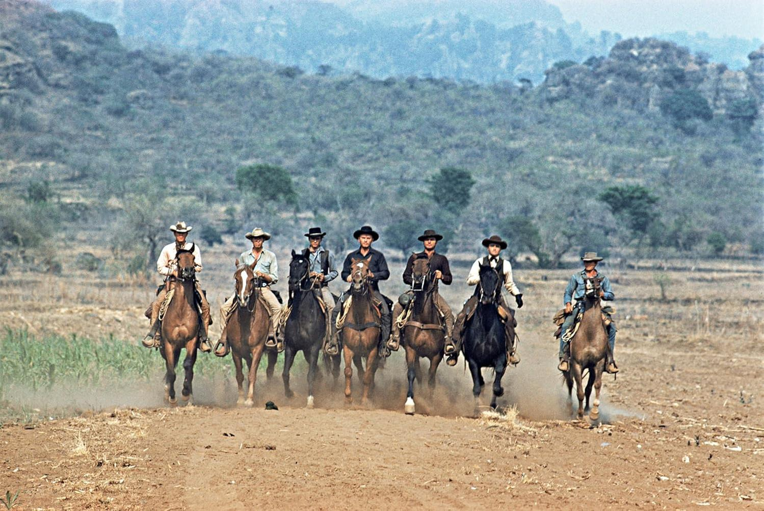 Sieben Cowboys auf Pferden reiten in Formation über ein trockenes, staubiges Feld. Berge sind im Hintergrund.