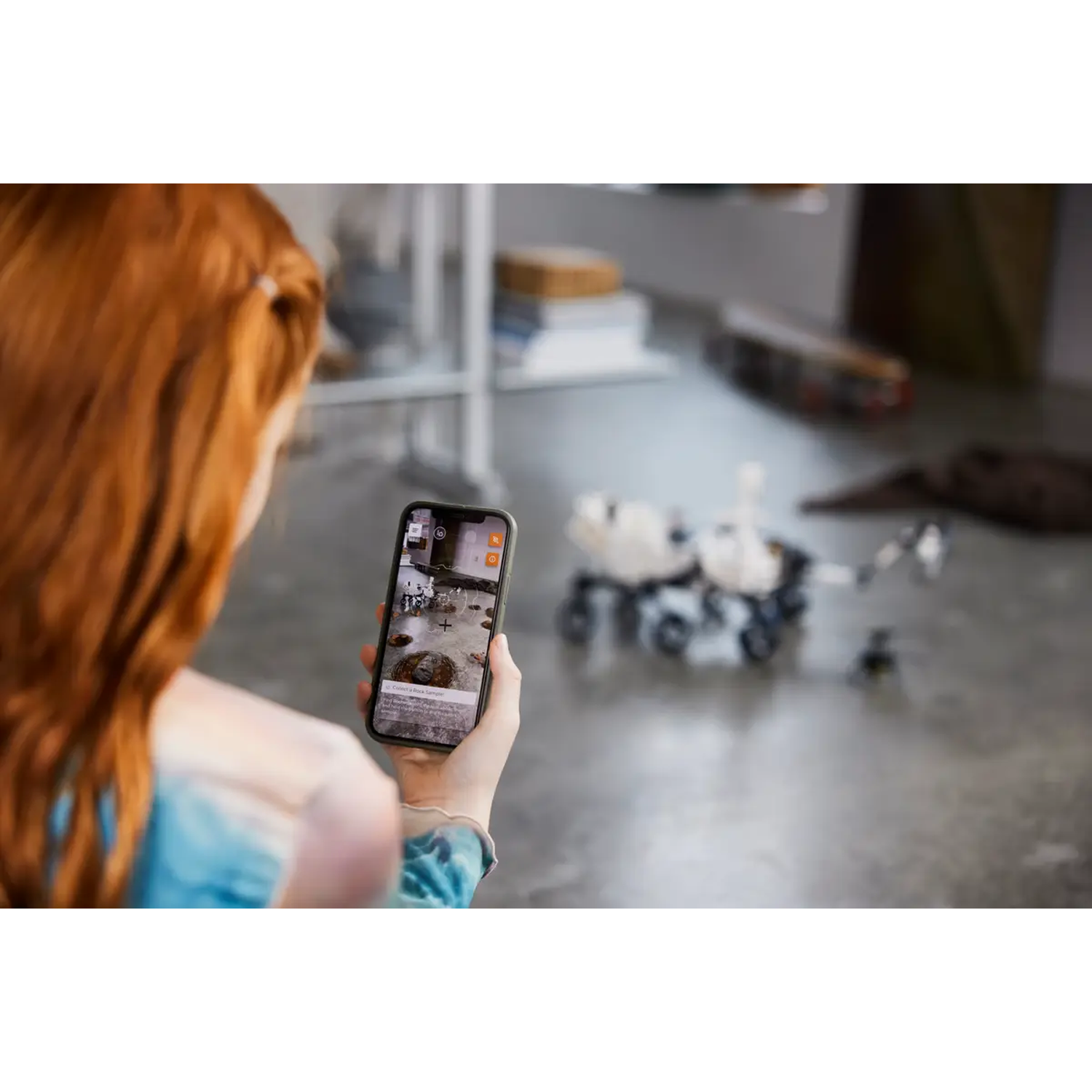 A person holds a phone showing a Mars rover model. The model is on the floor. The person has red hair and is wearing a blue shirt.