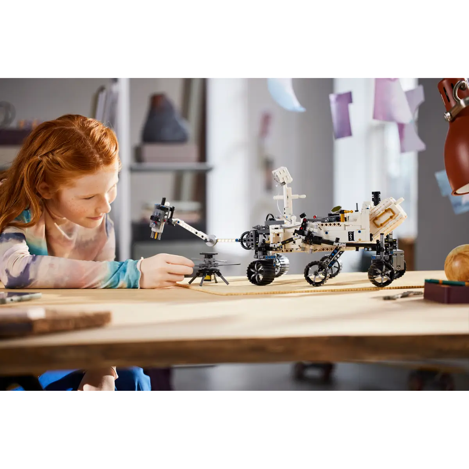 A girl with red hair is building a white and black Lego rover and helicopter on a wooden table.