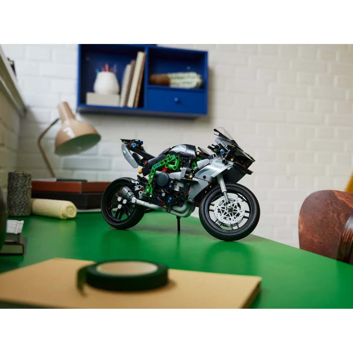 A toy motorcycle sits on a green desk. A roll of tape and a notepad are in the foreground. A shelf with books is in the background.