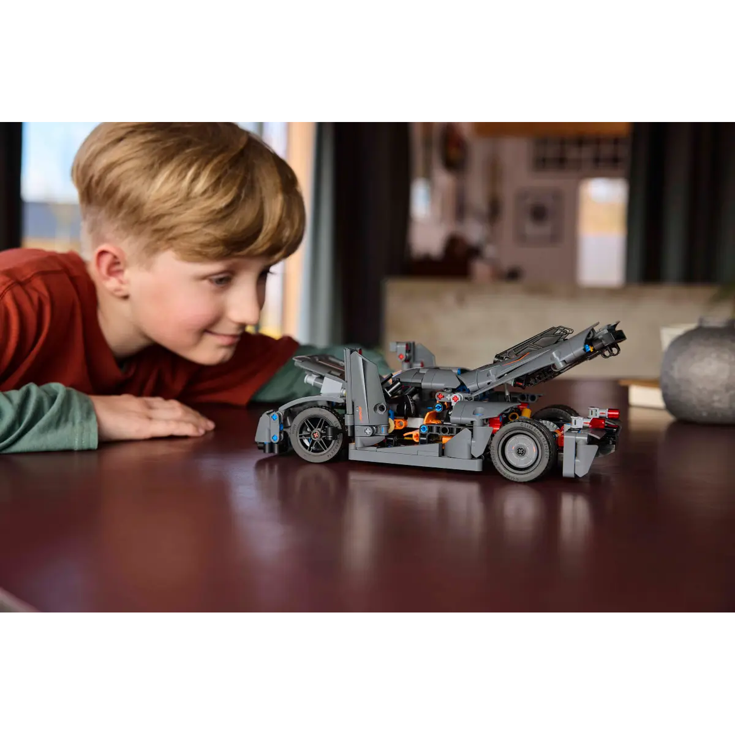 Een jongen bekijkt een grijze LEGO-auto op een bruine tafel. De auto heeft zwarte wielen en oranje accenten. De achtergrond is wazig.