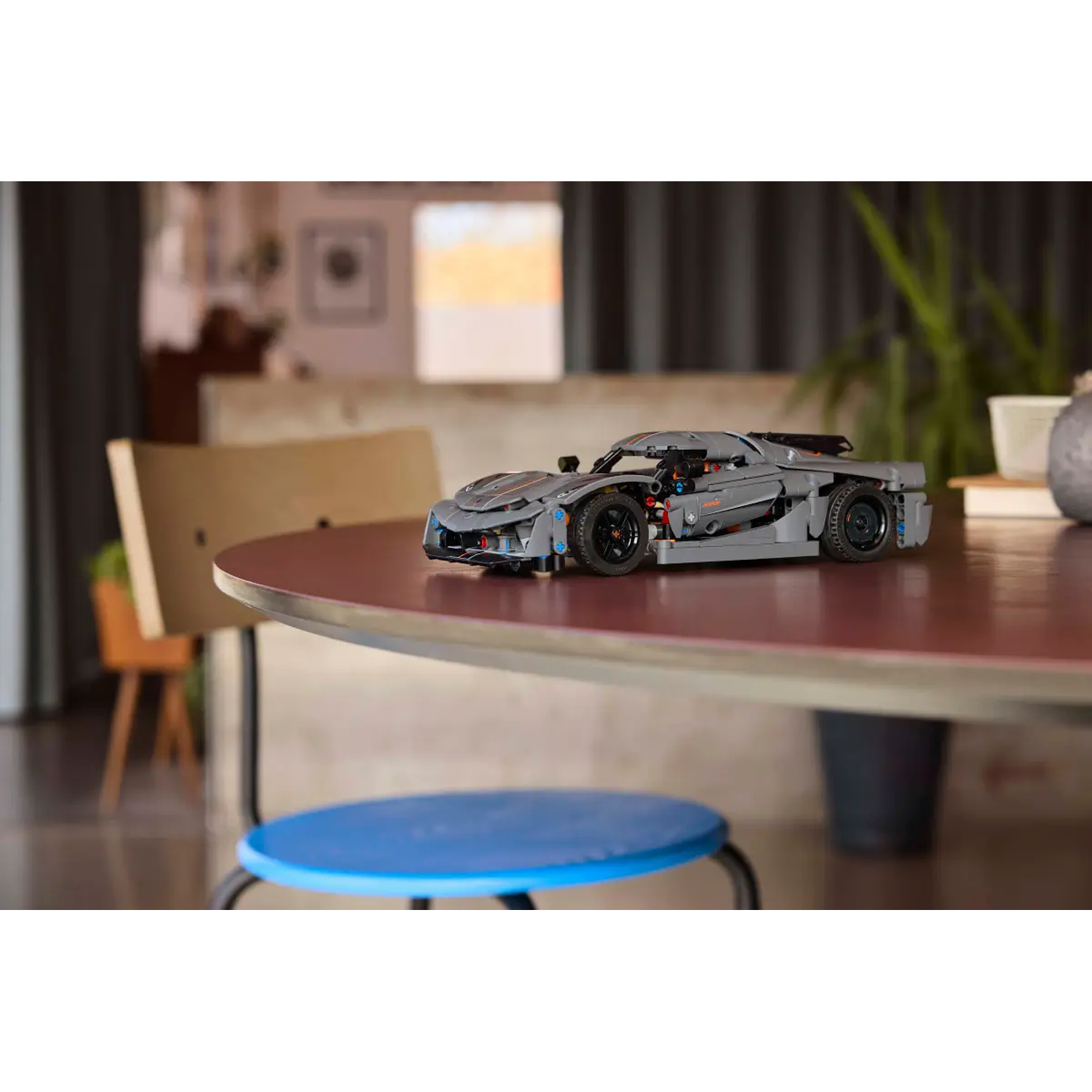 Een grijze Lego sportwagen staat op een ronde, rode tafel. Er staat een blauwe kruk voor. Achtergrond bevat een stoel en planten.